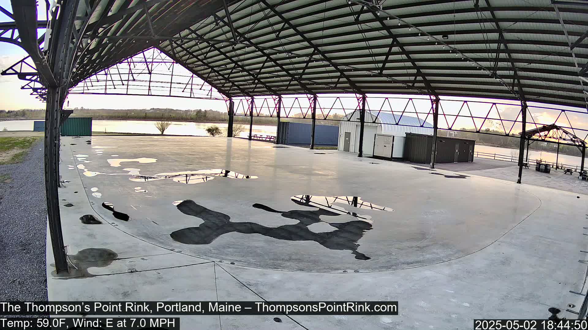 A large, empty concrete rink under a metal roof, with several puddles on the surface, overlooks a calm body of water under a partly cloudy sky.