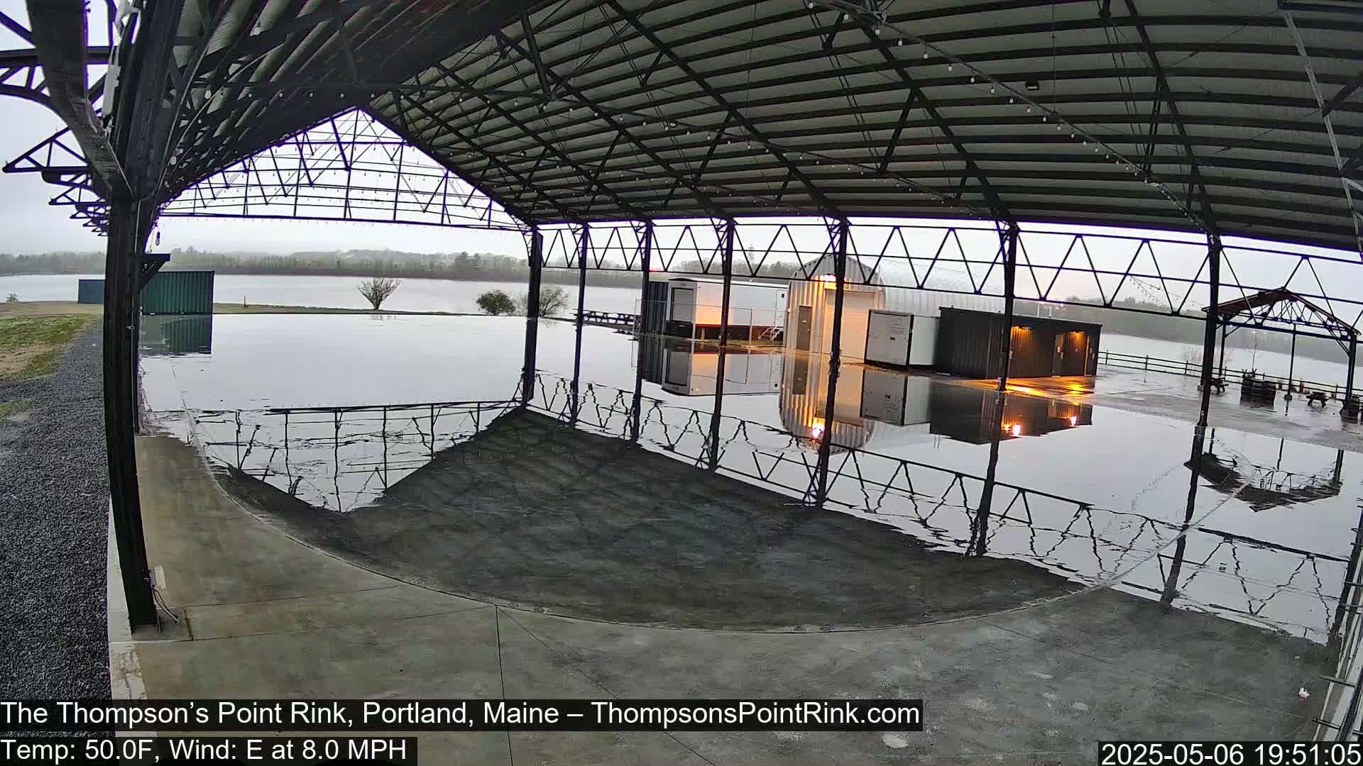 Under an overcast sky, a flooded outdoor rink sits under a metal-framed structure with a calm body of water visible in the background.