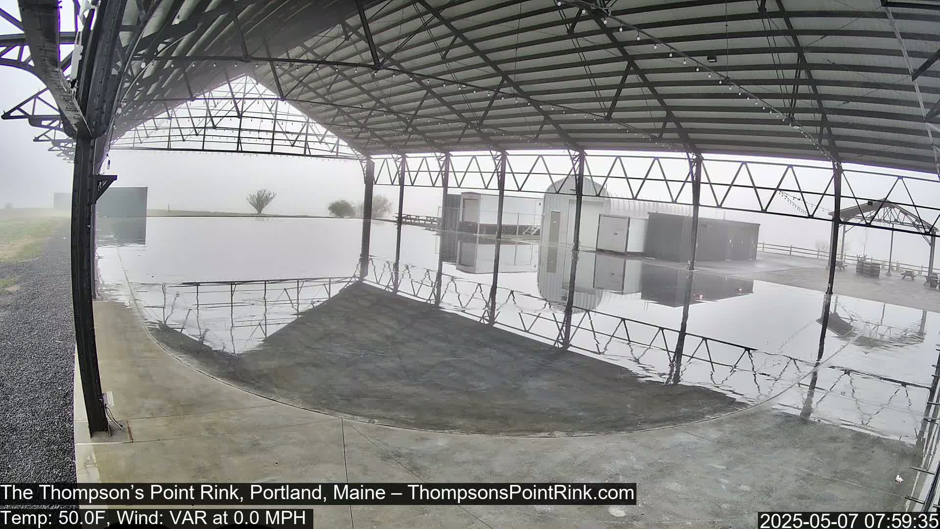 A fog-shrouded outdoor rink sits under a metal-framed shelter, its surface wet and reflecting the structure above.
