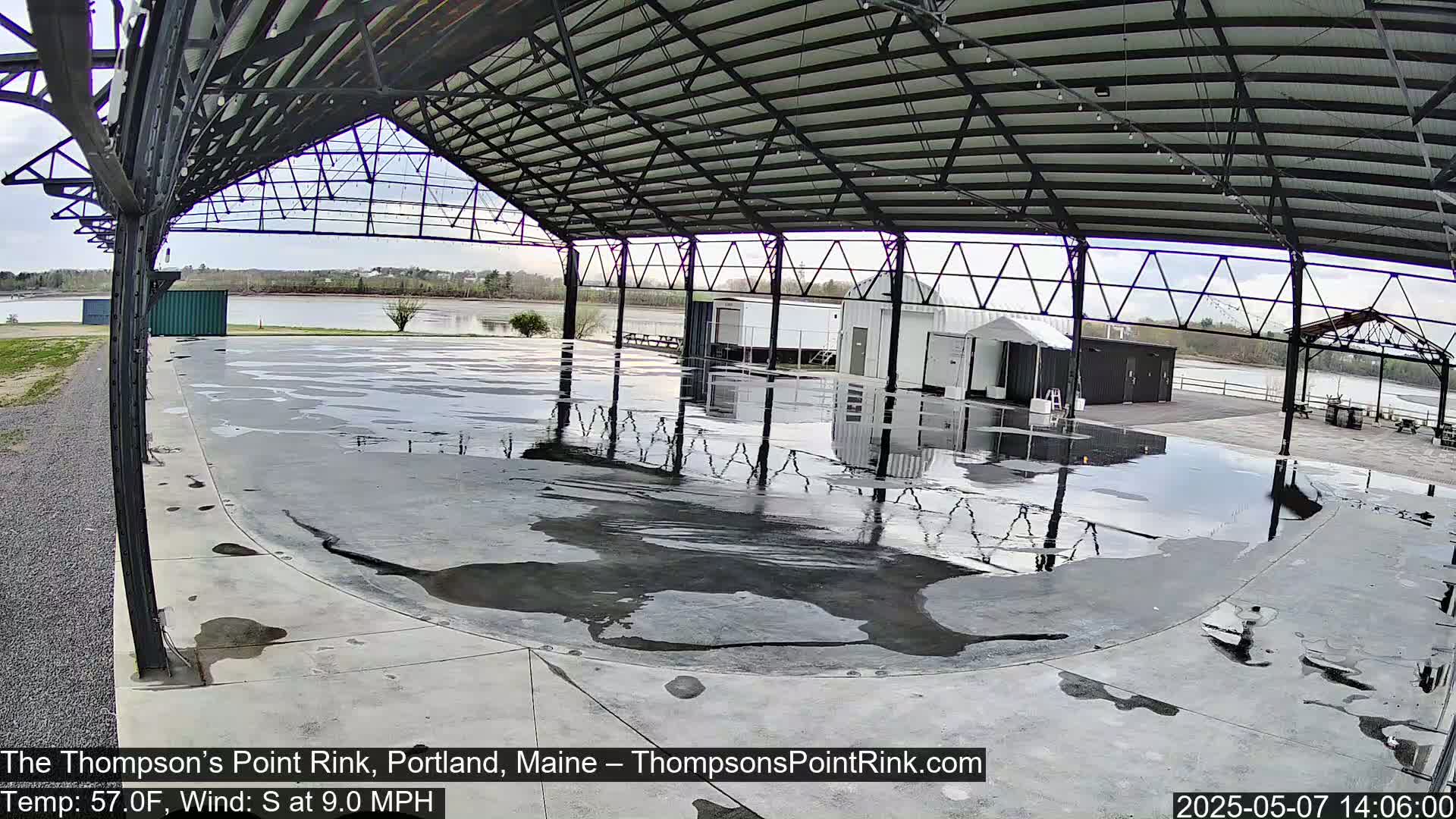 A wet, open-air rink under a metal structure overlooks a calm body of water on a partly cloudy day with a south wind.