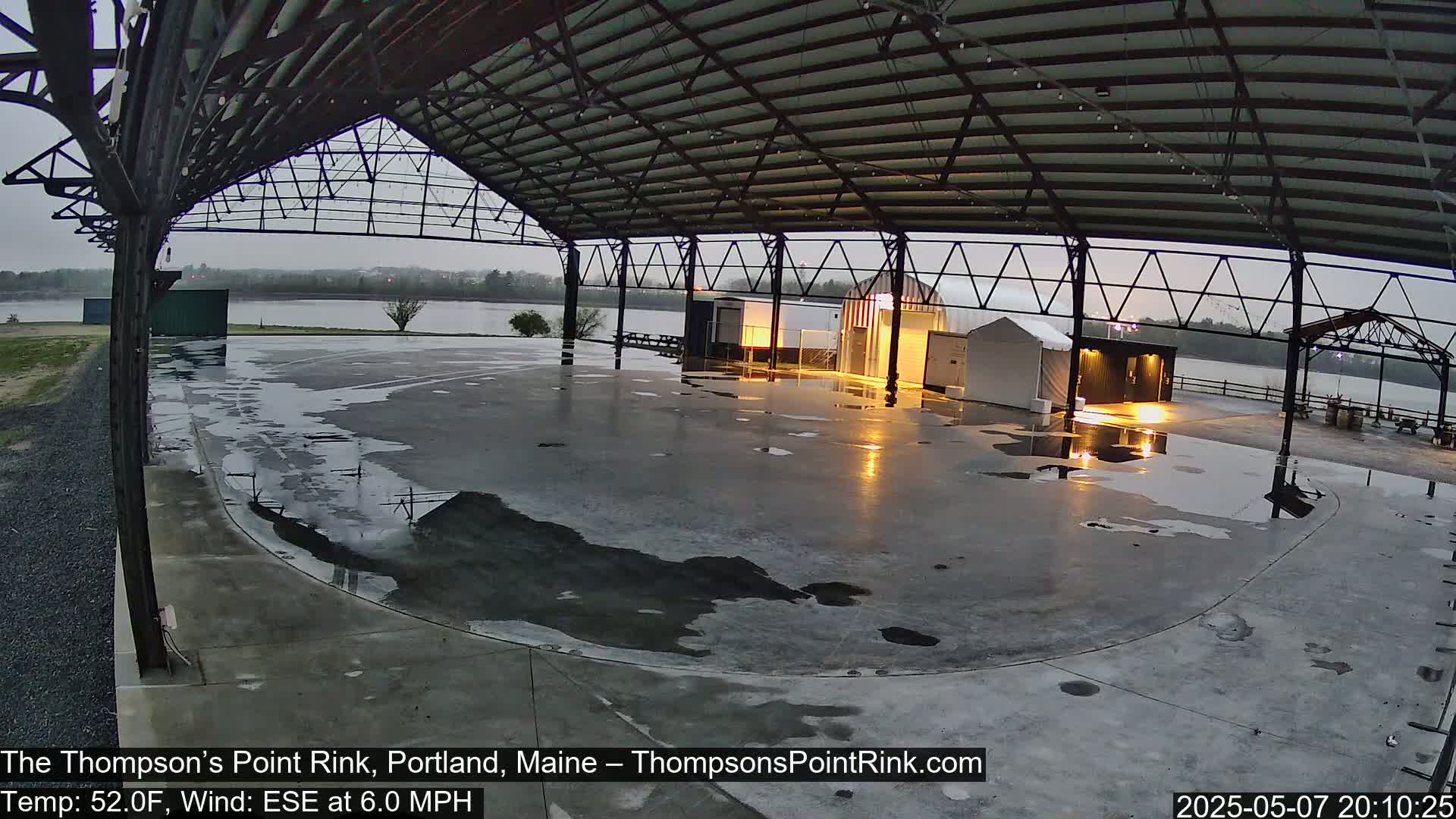 A wet, open-air rink under an overcast sky is seen with puddles on the ground and a calm body of water visible in the background.