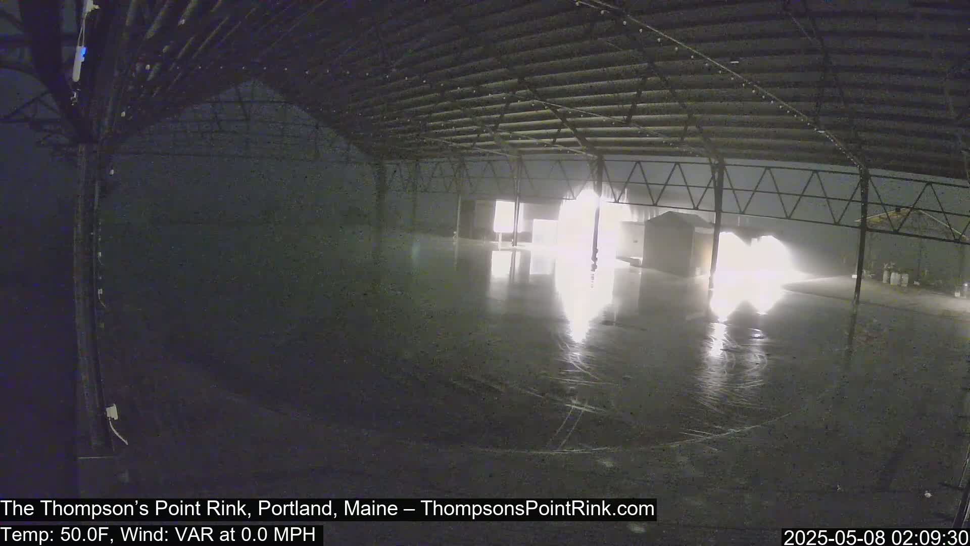A brightly lit, empty, wet indoor rink is seen at night under a dark, possibly overcast sky.