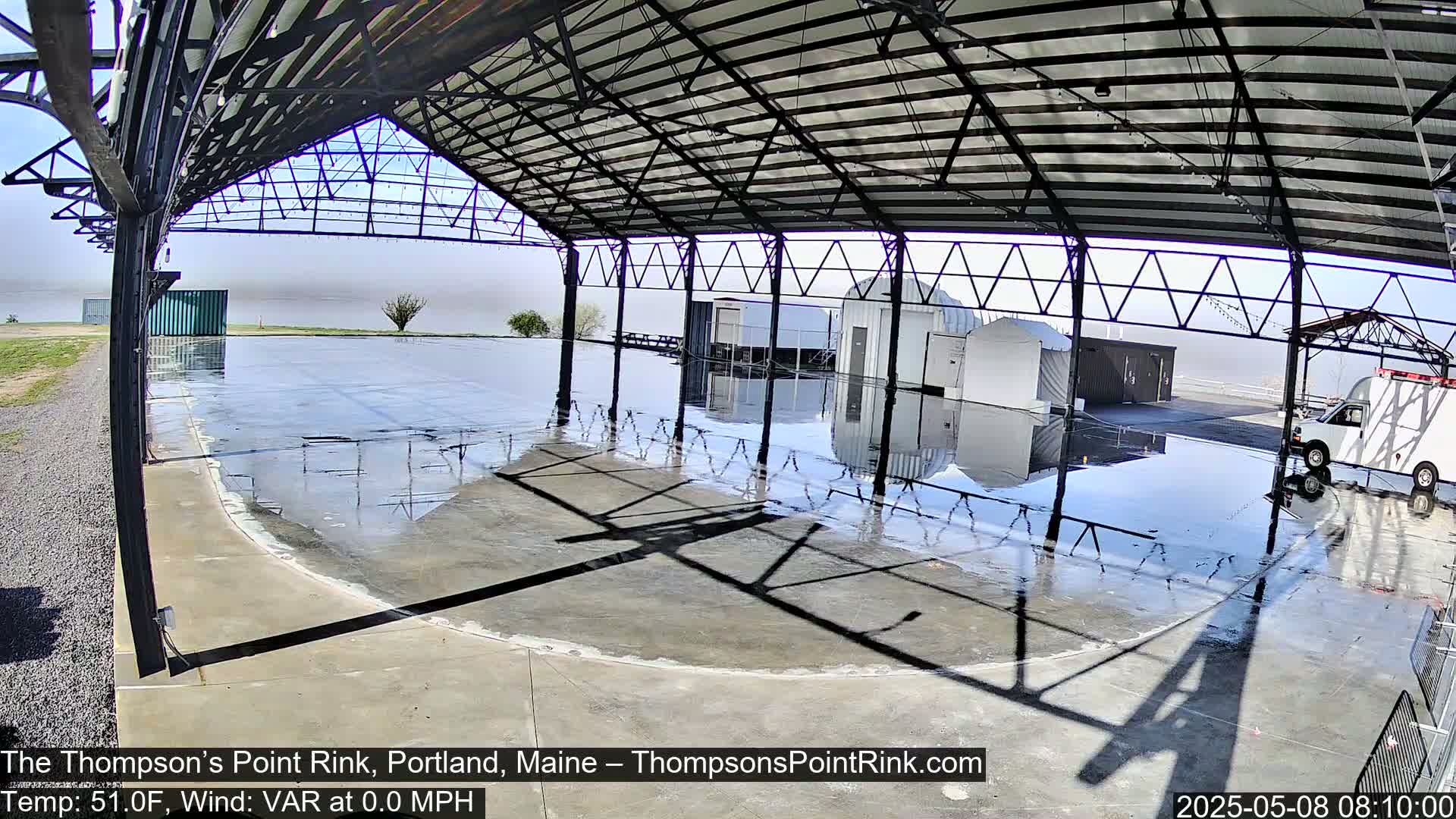 A wet, mostly empty concrete rink sits under a metal-framed shelter on a hazy day.