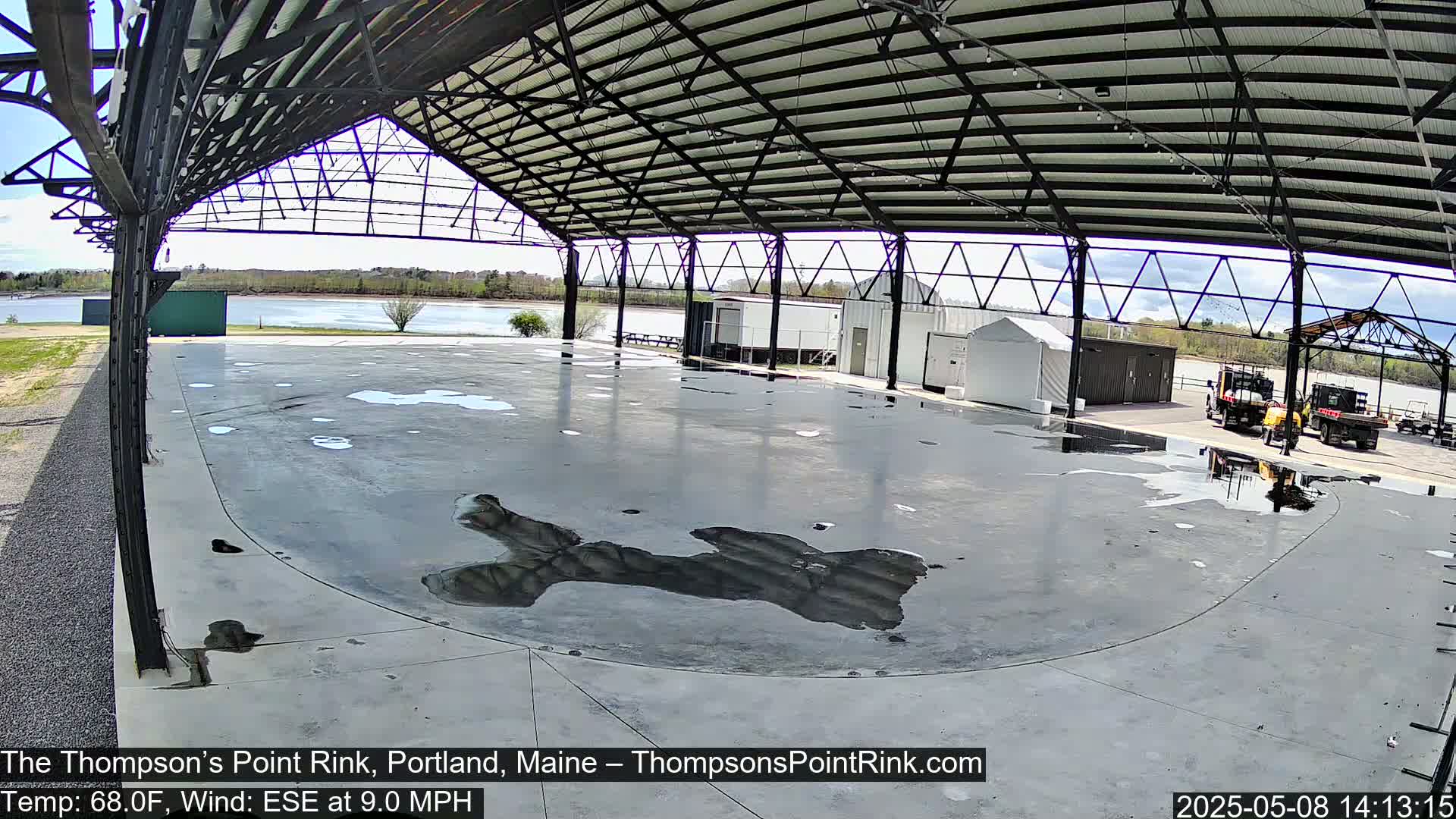 An empty, wet rink sits under a metal-framed roof, overlooking a calm body of water on a partly sunny day.