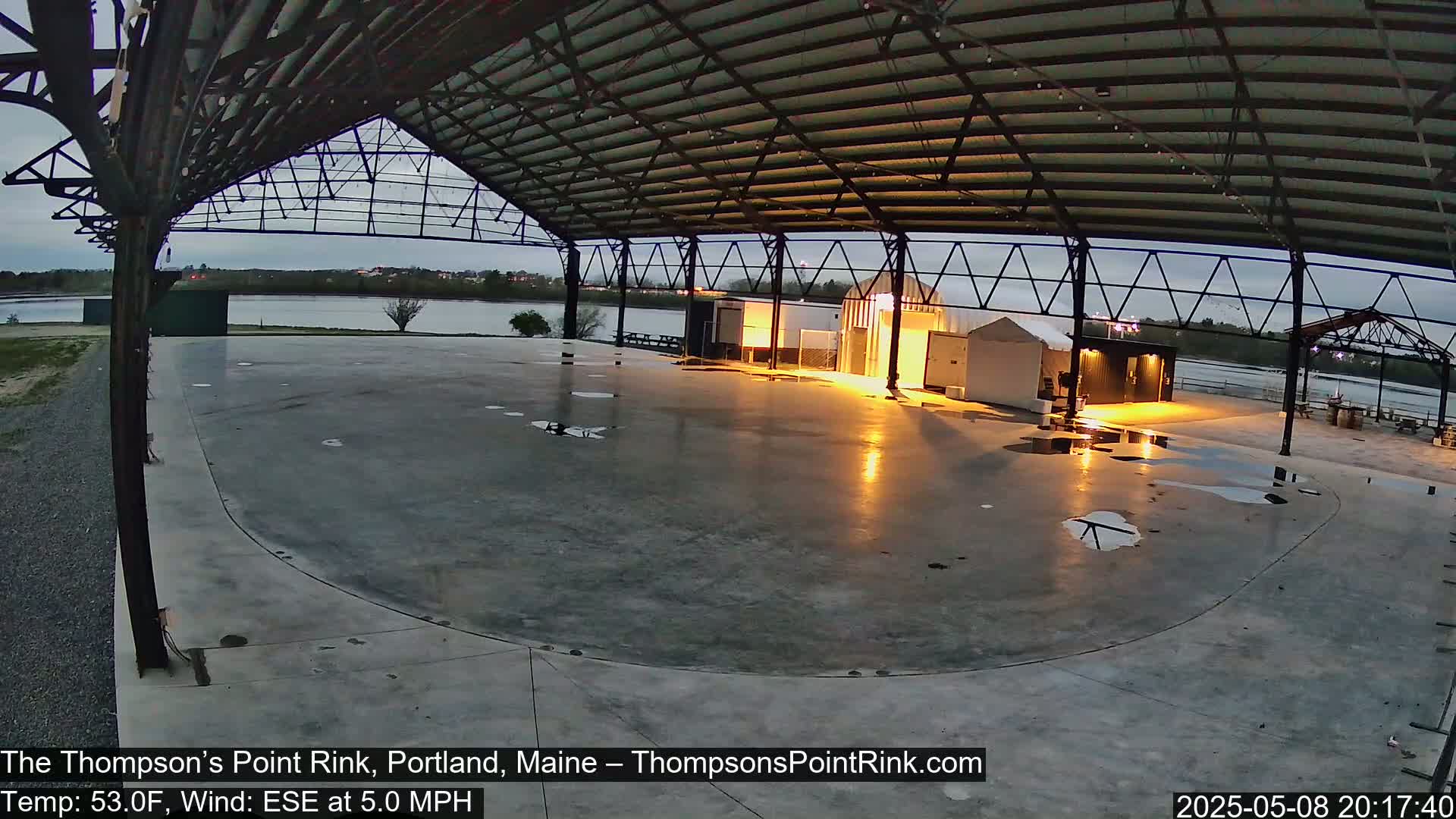 A wet, empty concrete rink is under a metal roof structure at dusk, with a calm body of water visible in the background.