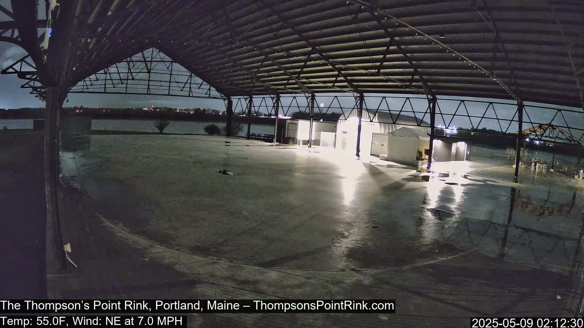 A wet, empty paved area under a large metal structure at night, with a body of water visible in the background under a dark, overcast sky.