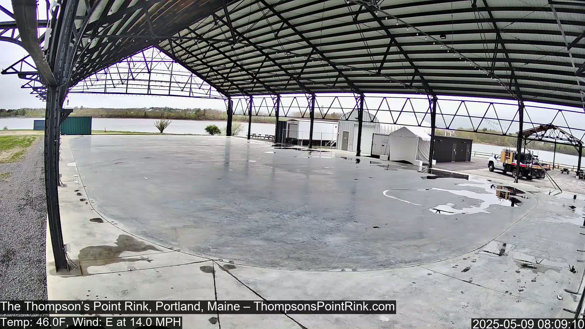 A wet, empty rink under a metal-framed roof overlooks a calm body of water on an overcast day.