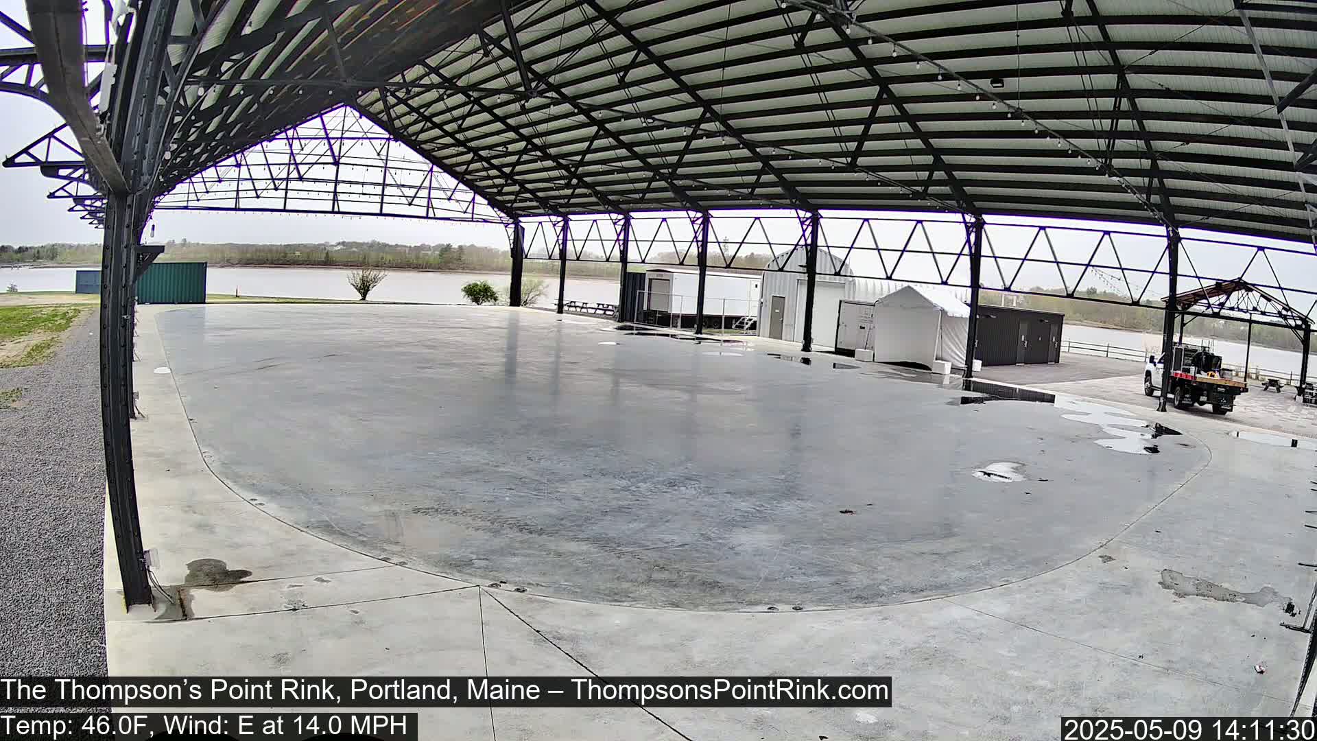 A wet, empty concrete rink is situated under a metal-framed open-air structure overlooking a calm river on a partly cloudy day.