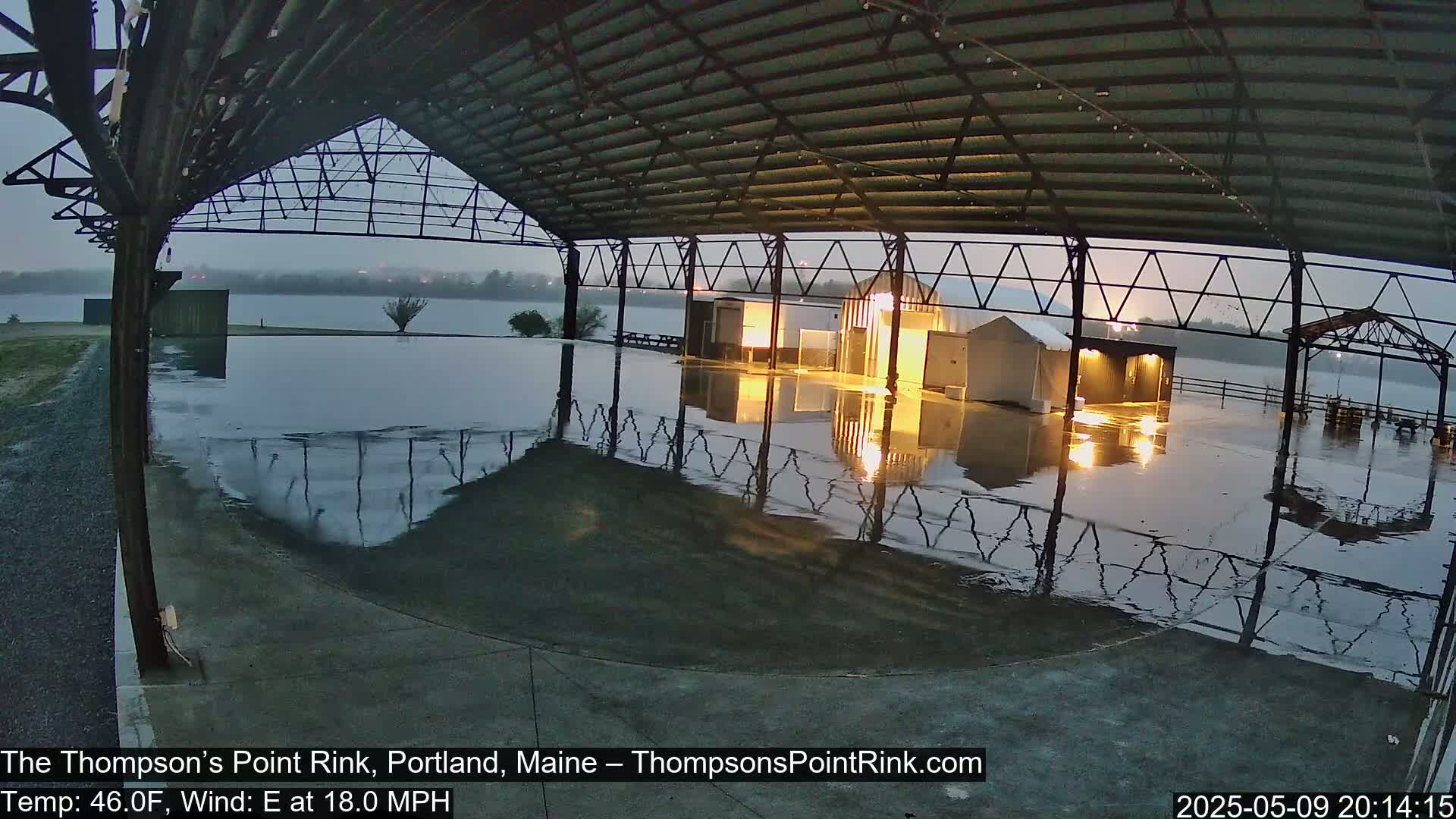 A flooded rink under a metal-framed shelter is viewed from inside, with an overcast sky and an 18 mph east wind.