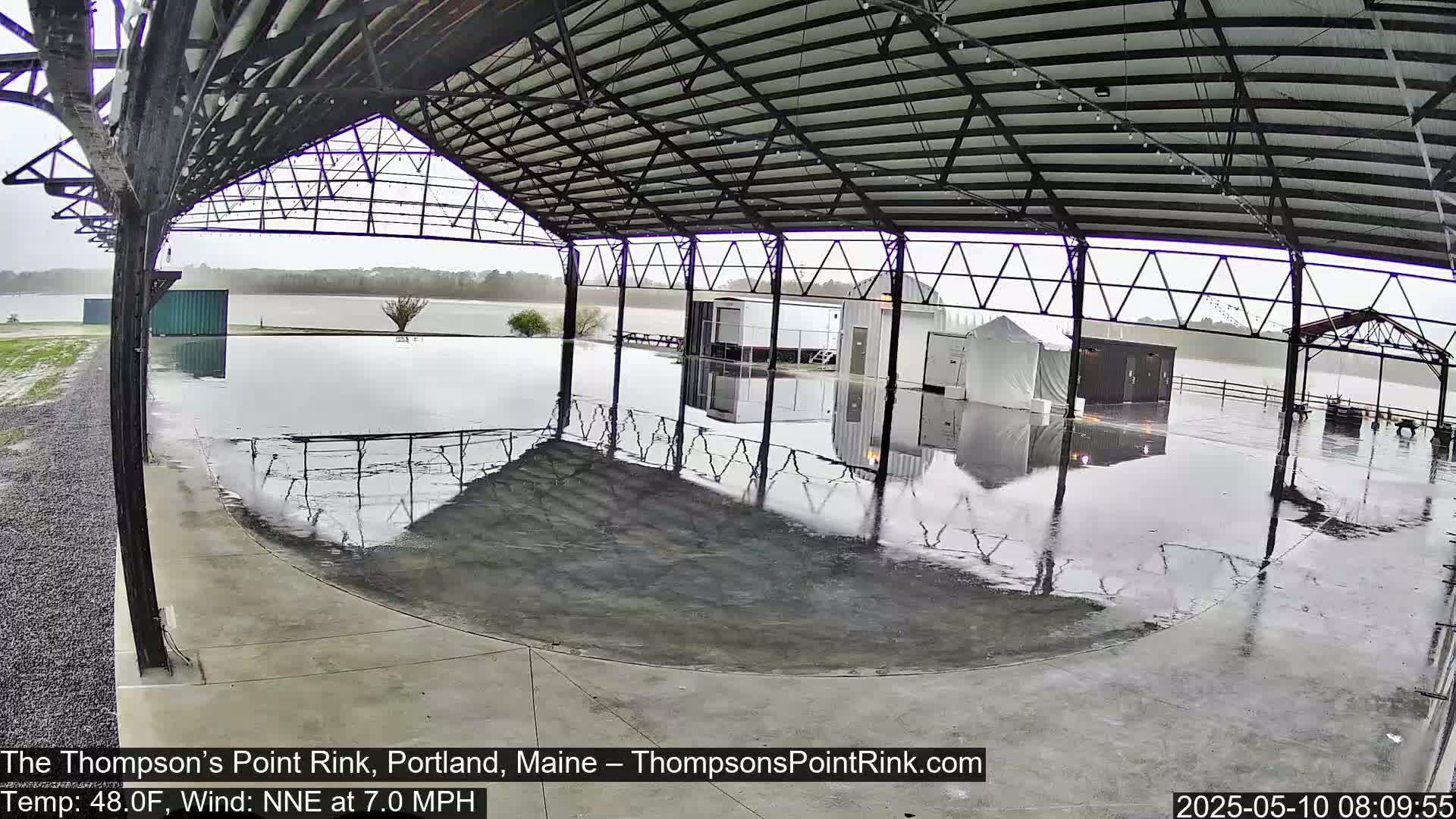 A flooded open-air rink under a metal roof is seen during rainy weather.