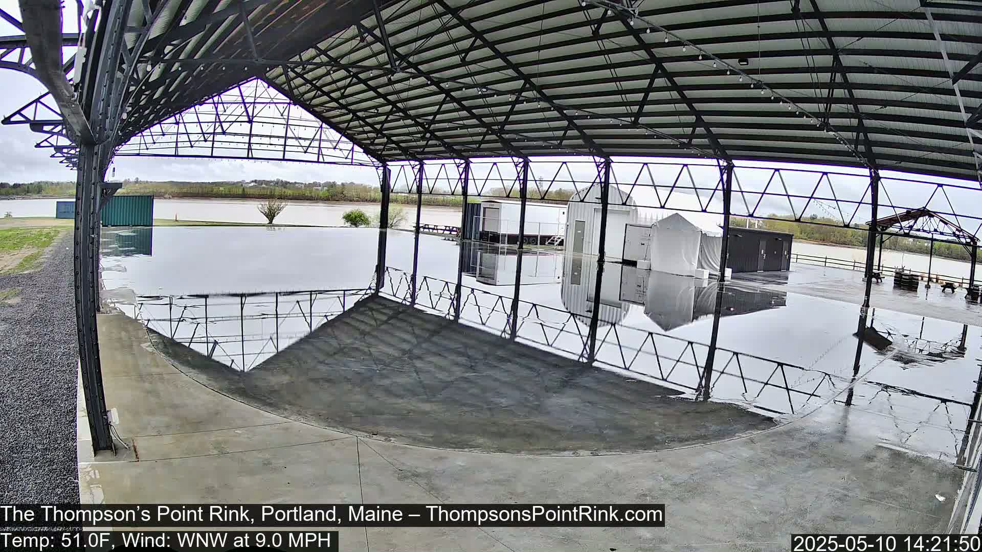 A wet, open-air rink under a metal structure overlooks a calm body of water on an overcast day.