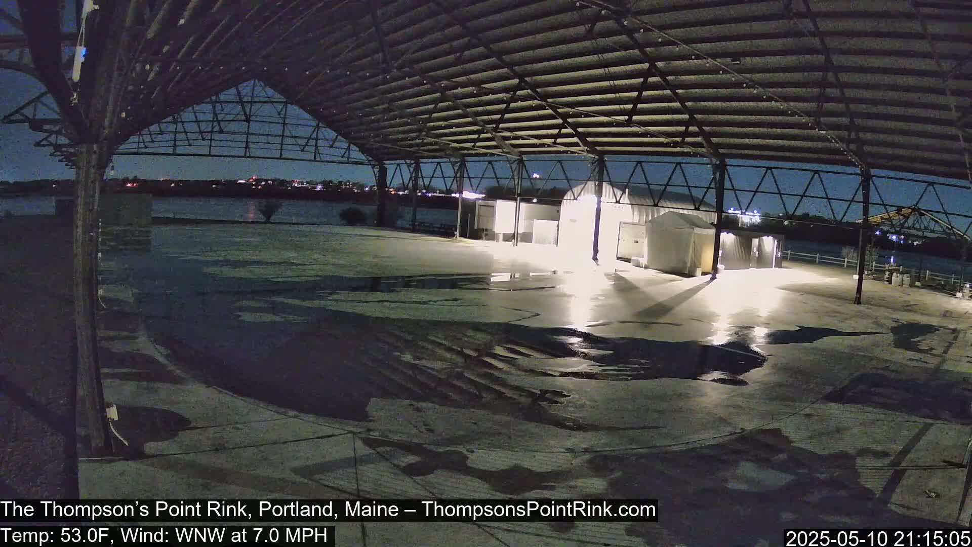 An empty, wet rink under a metal roof at night, with a calm, dark body of water visible in the distance.