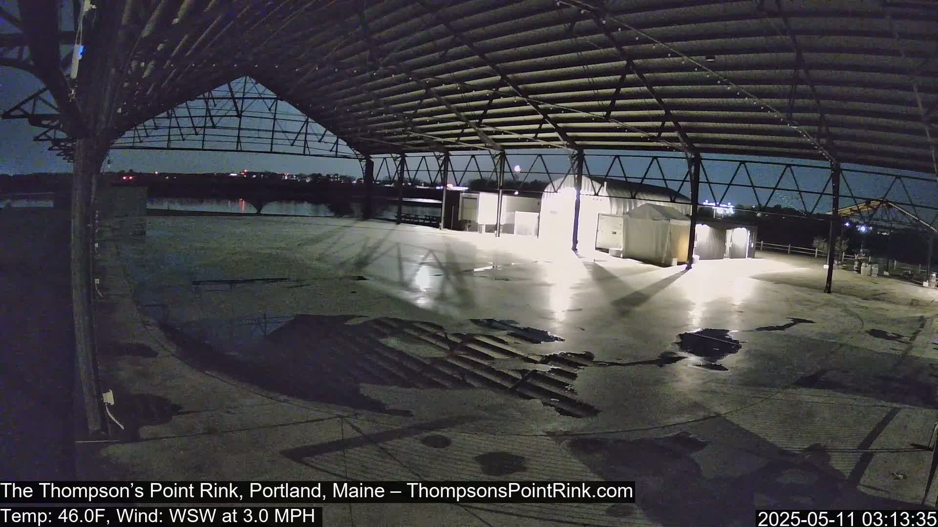An empty, open-air rink at night under a metal roof, with puddles on the concrete floor and a calm, clear sky.