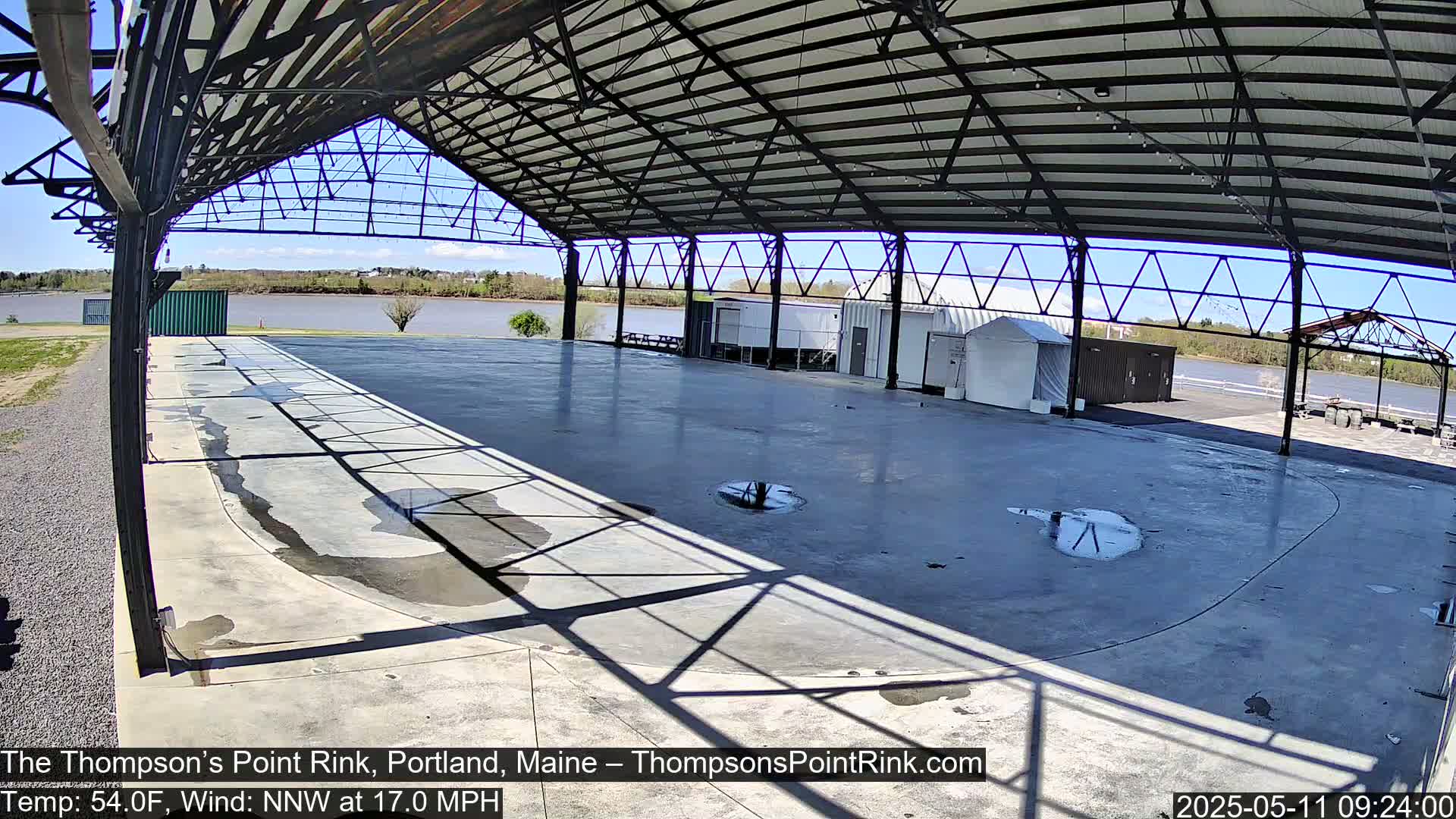 A large, empty, open-air concrete rink with a metal roof structure is situated beside a river under a sunny sky.