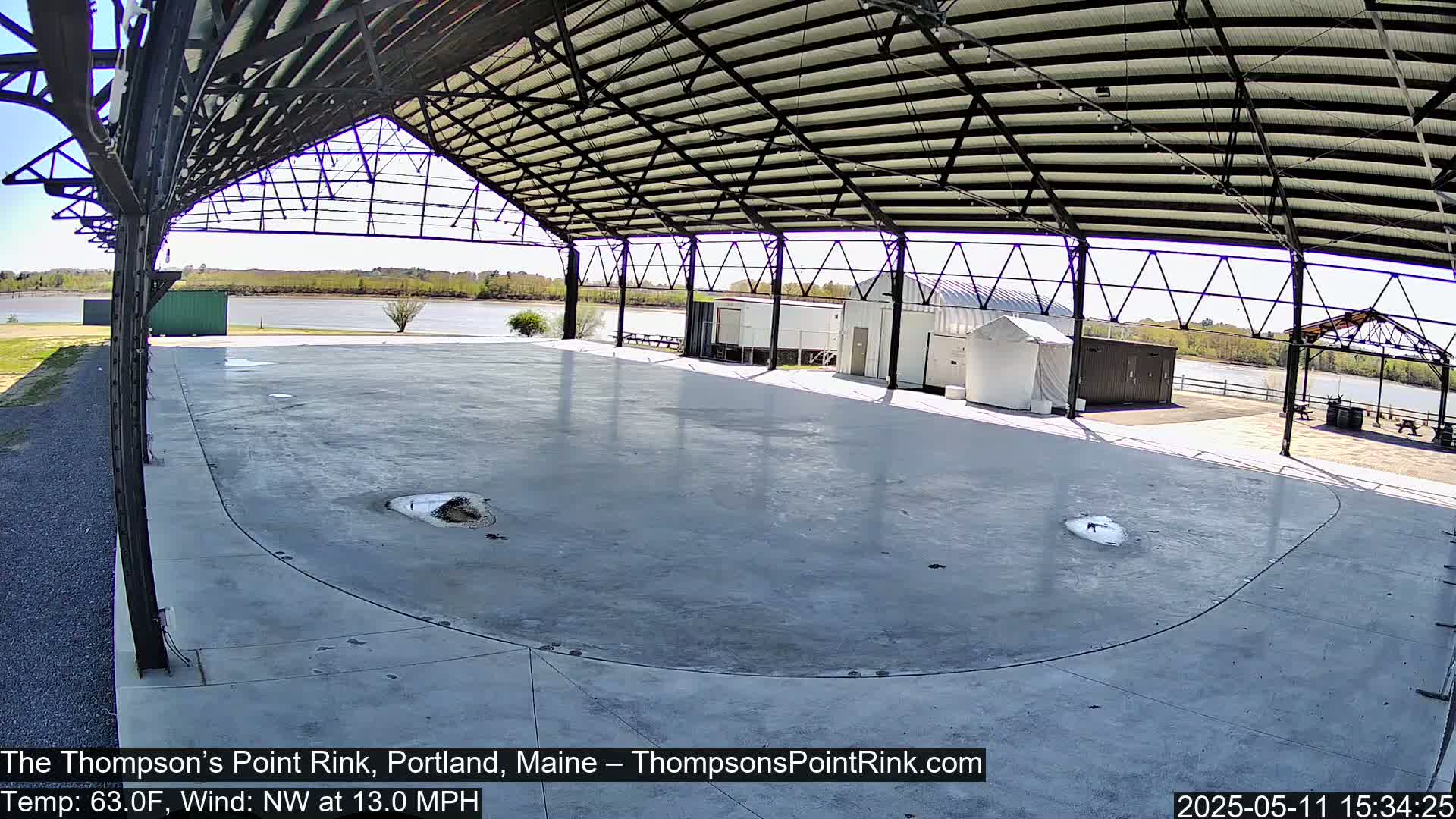 A large, empty, concrete rink sits under a metal-framed shelter overlooking a calm body of water on a sunny day with a northwest wind.