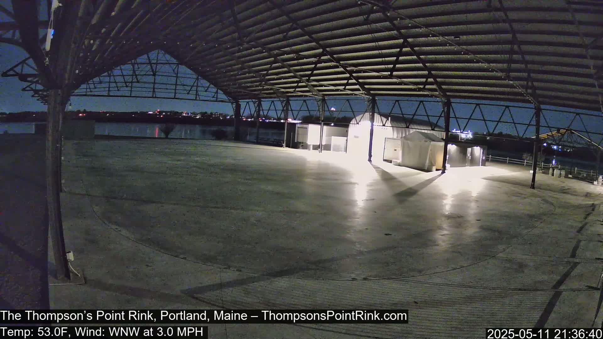 An empty, paved rink sits under a metal structure at night, overlooking a calm body of water under a clear sky.