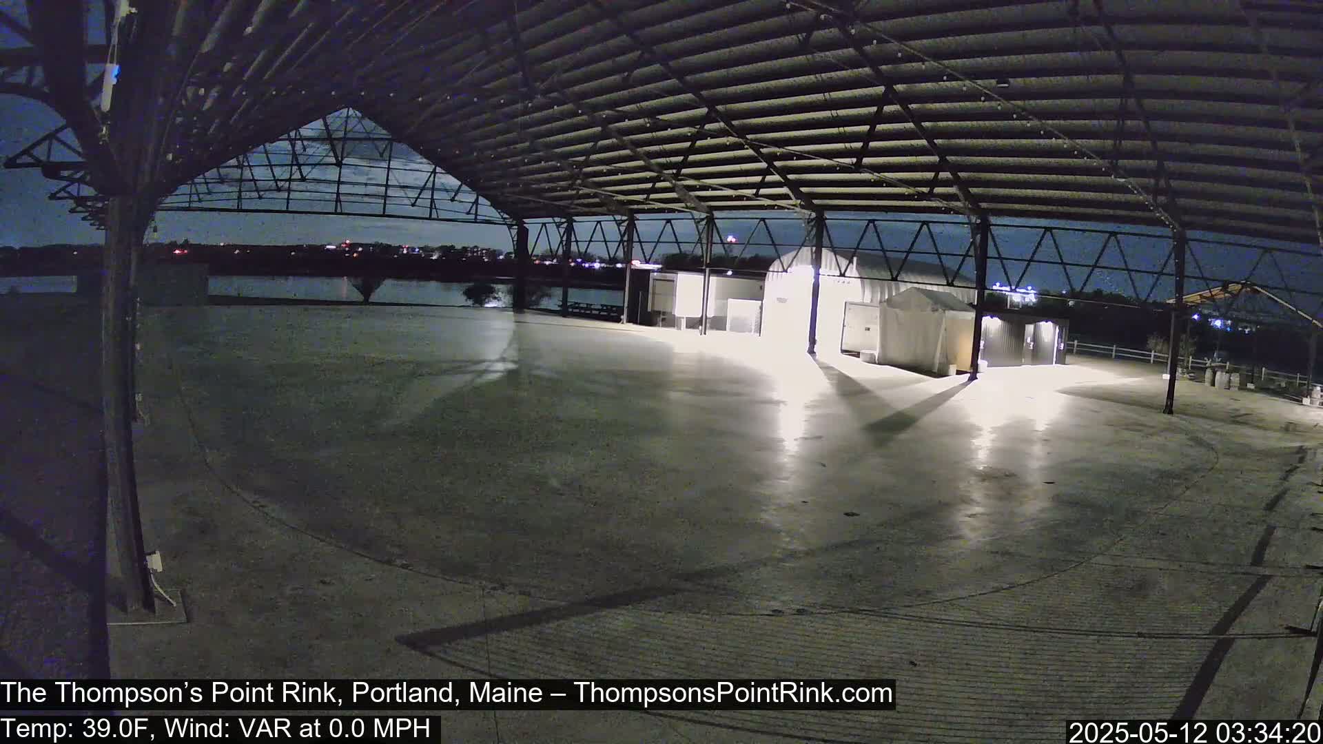 An empty, open-air rink under a metal roof at night, illuminated by lights, with calm weather.