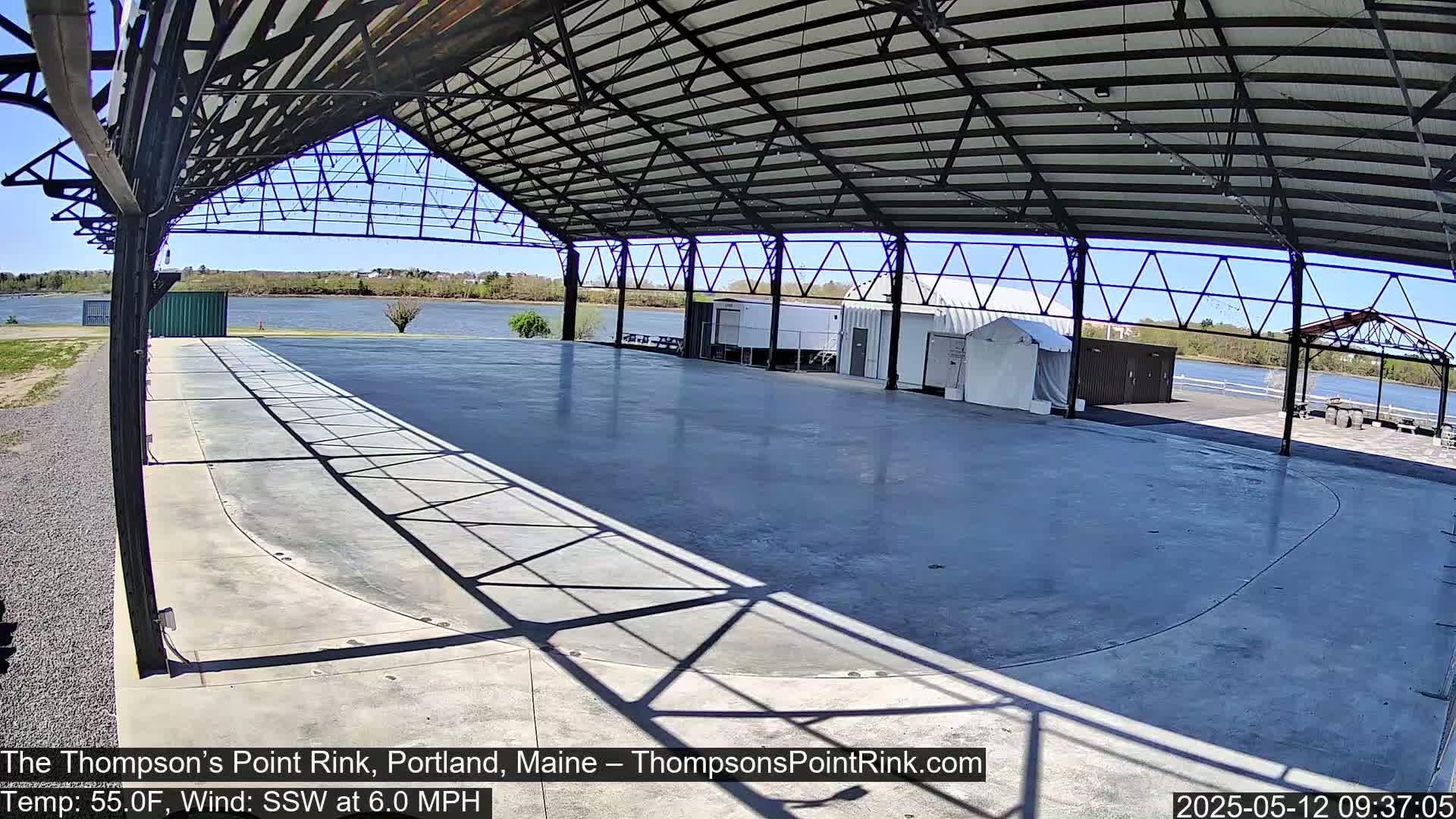 A large, empty, open-air rink under a metal structure overlooks a calm body of water on a sunny day with a light south-southwesterly wind.