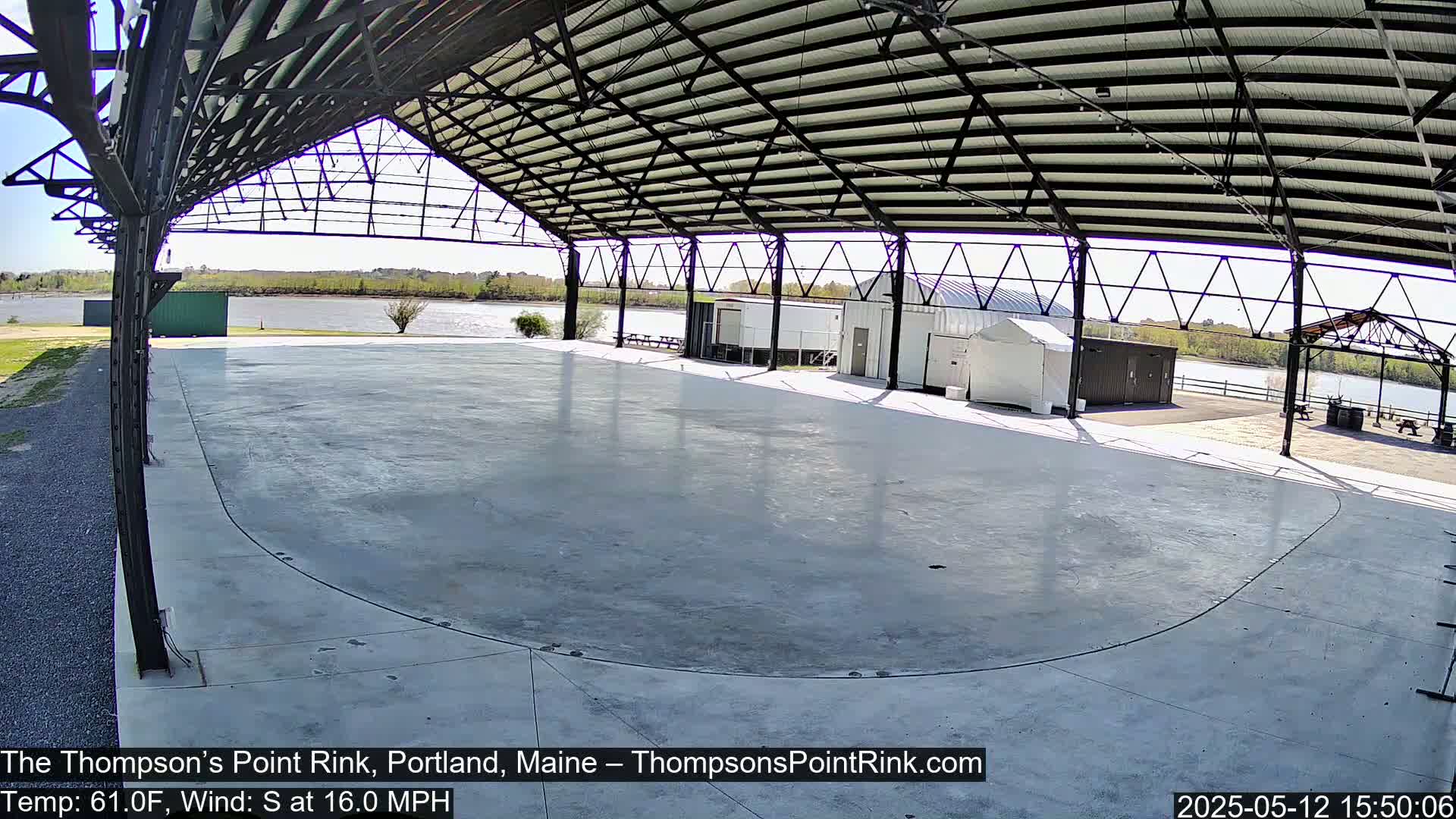 A large, empty, concrete rink under a metal-framed shelter overlooks a calm body of water on a sunny day with a south wind.