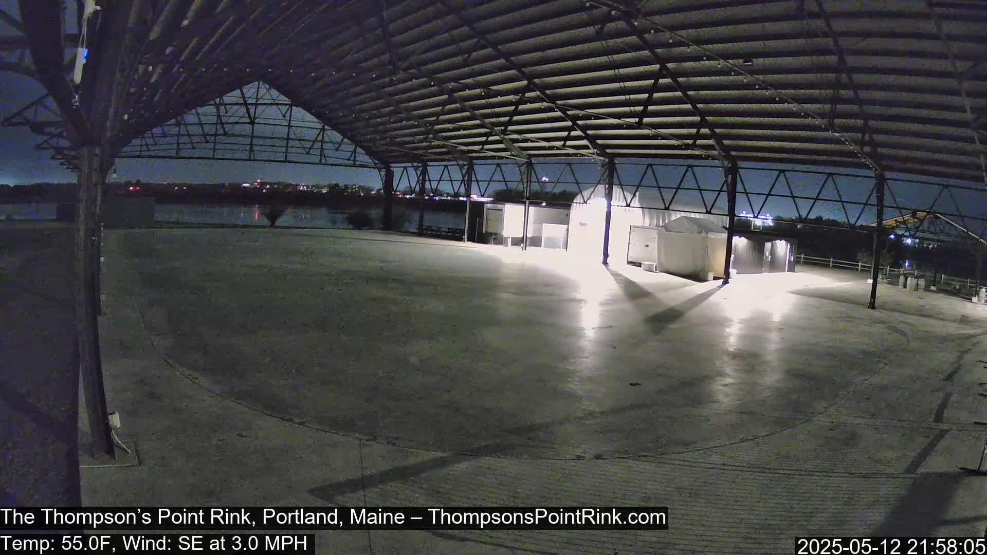 An empty, open-air rink under a metal roof at night, illuminated by artificial light, with a calm body of water visible in the distance.