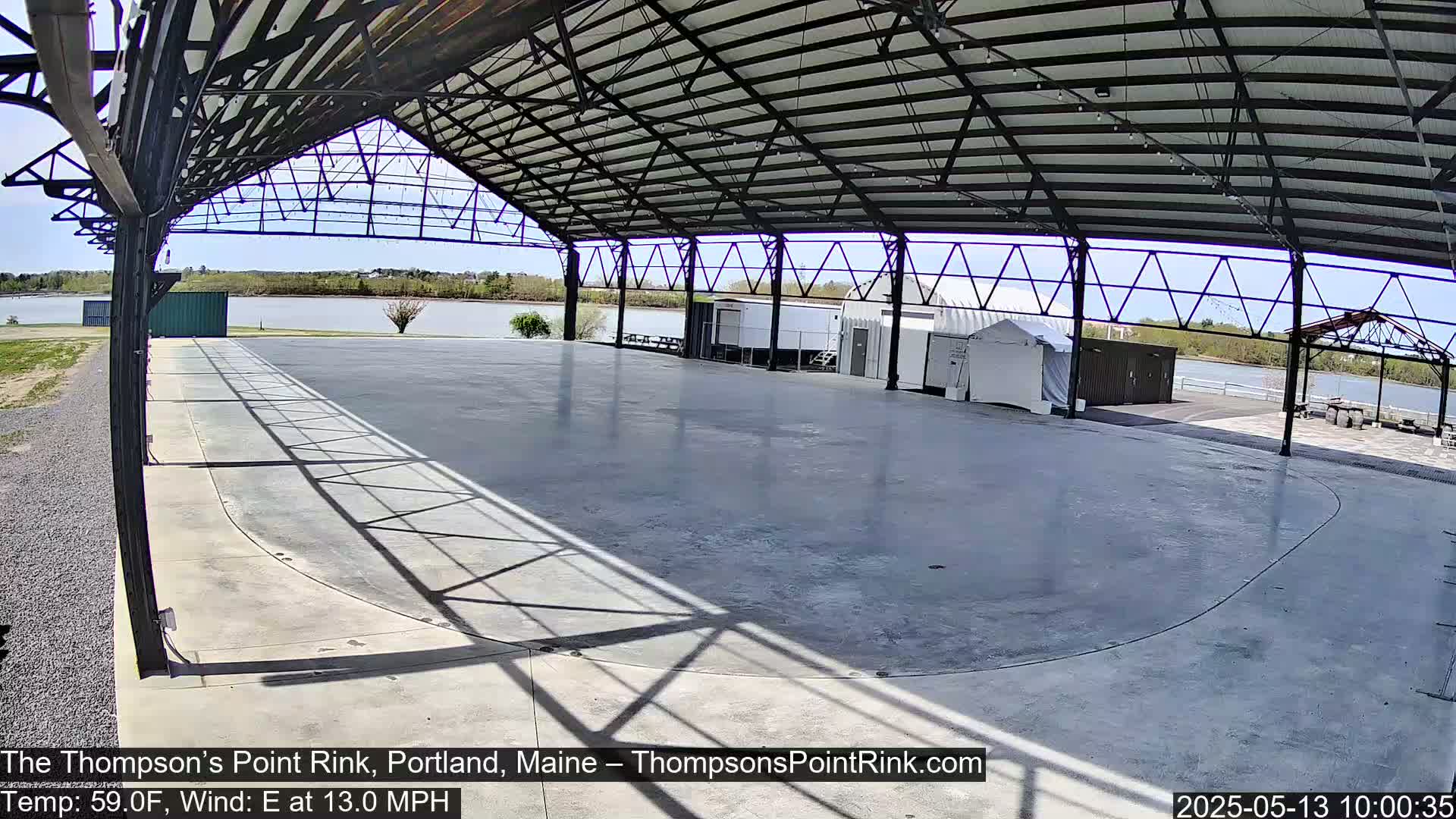 A large, empty, paved rink is enclosed by a metal-framed structure overlooking a calm body of water under a sunny sky.