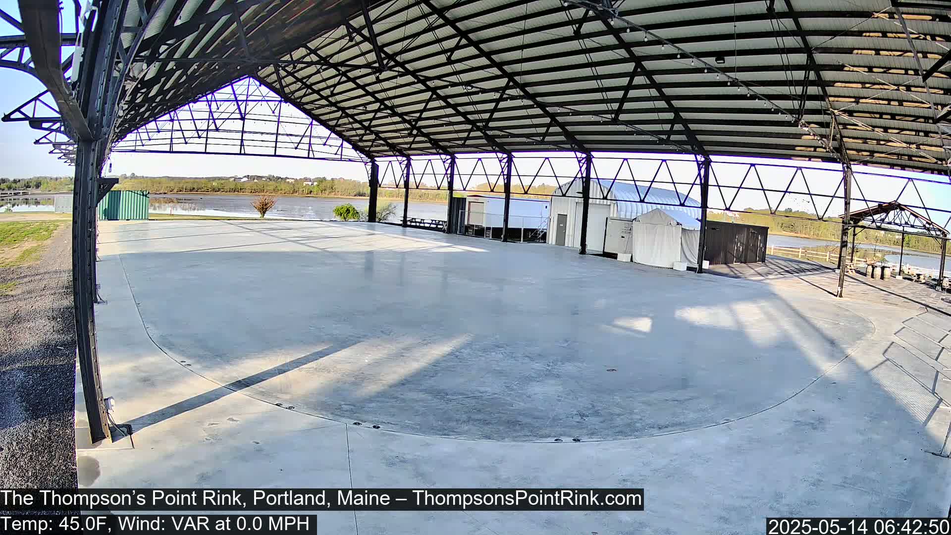 A large empty concrete rink is enclosed by a metal-framed structure, overlooking a calm body of water under a mostly clear sky.