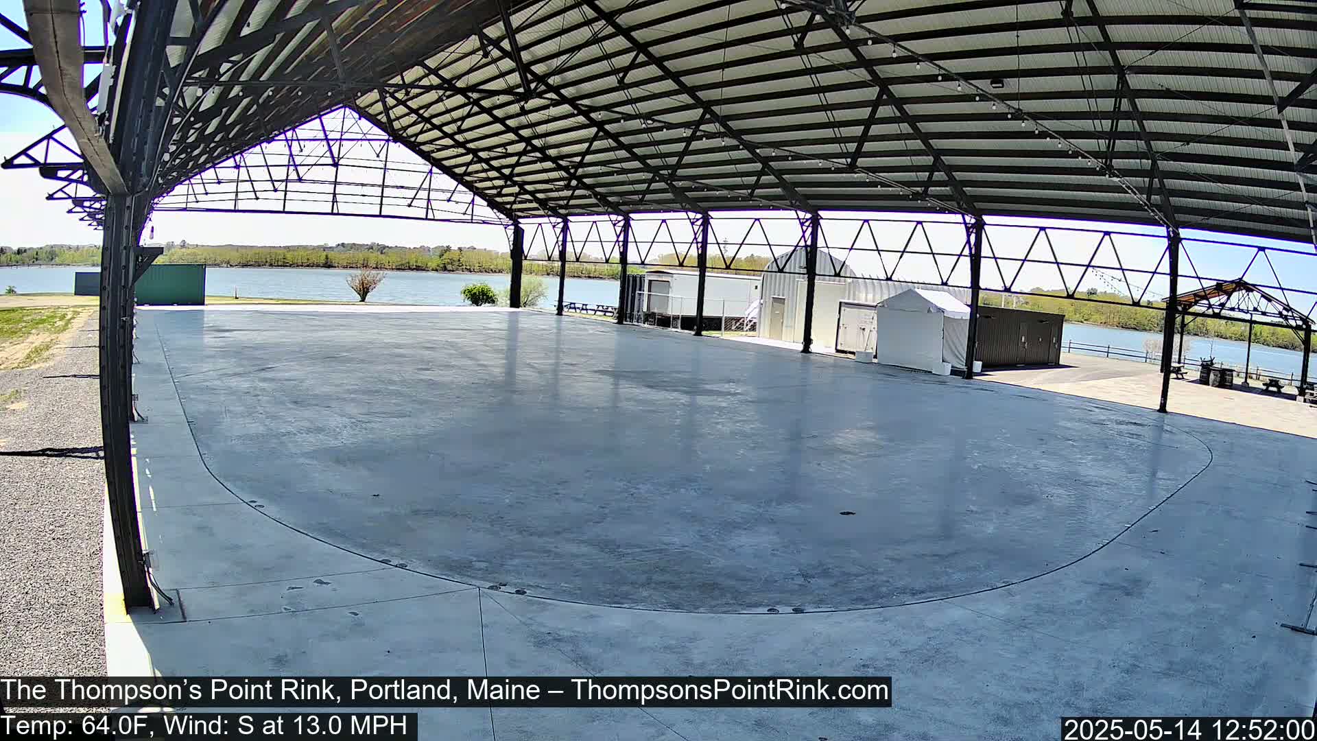 A large, empty concrete rink is enclosed under a metal-framed structure, overlooking a calm body of water on a sunny day with a south wind.