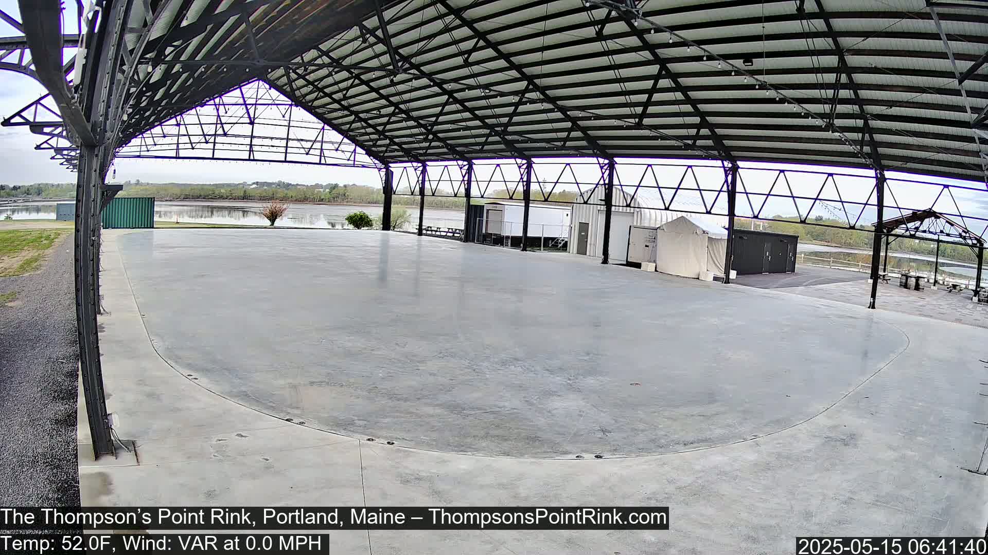 An empty, curved concrete rink sits under a metal-framed pavilion overlooking calm water on a partly cloudy day.