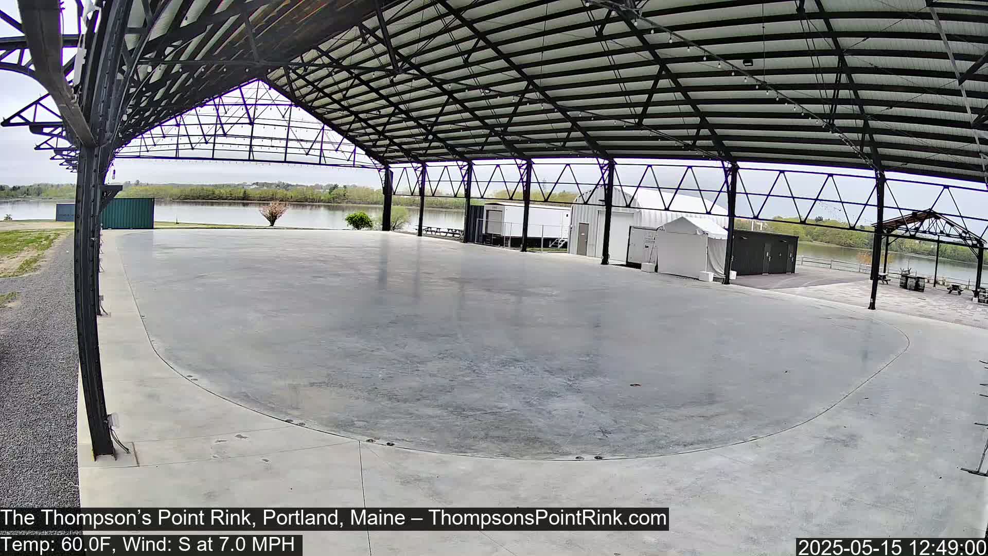 A large, empty concrete rink sits under a metal-framed roof, overlooking a calm body of water on a partly cloudy day.