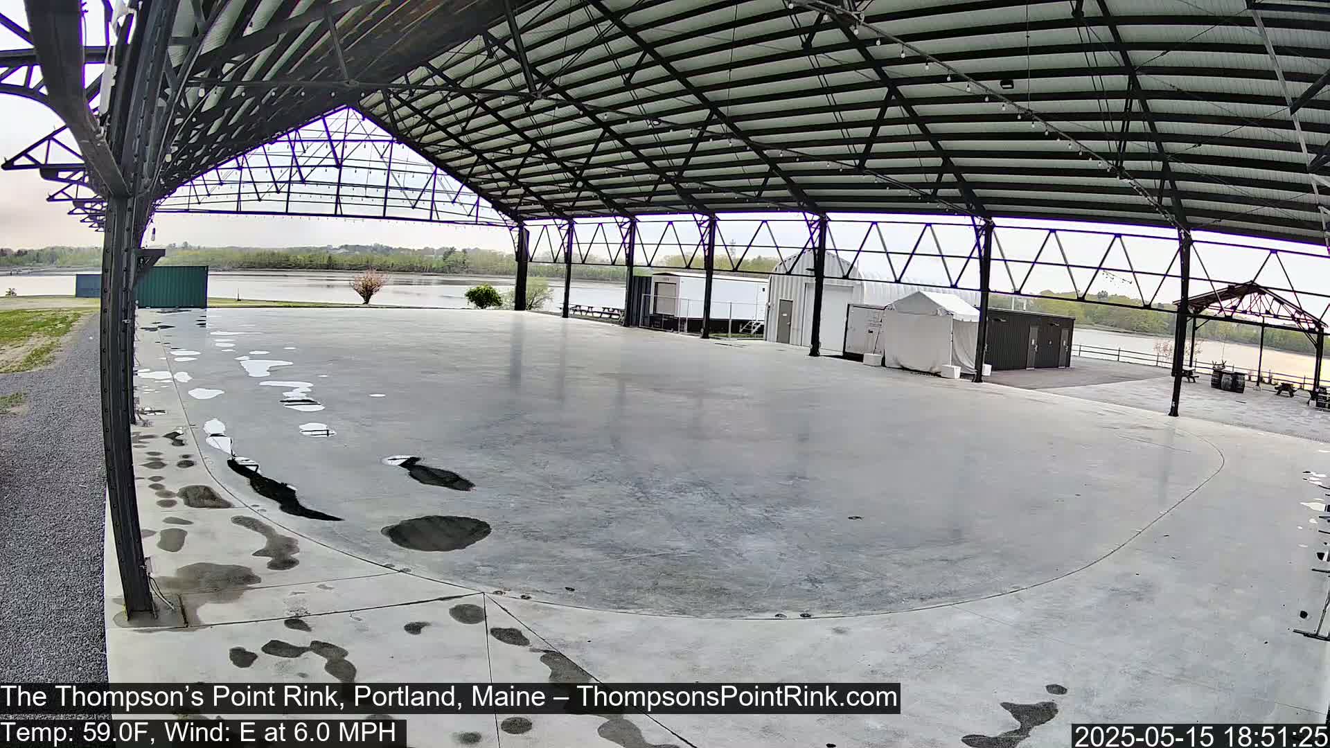 A large, empty concrete rink under a metal-framed covered structure overlooks a calm body of water on an overcast day.