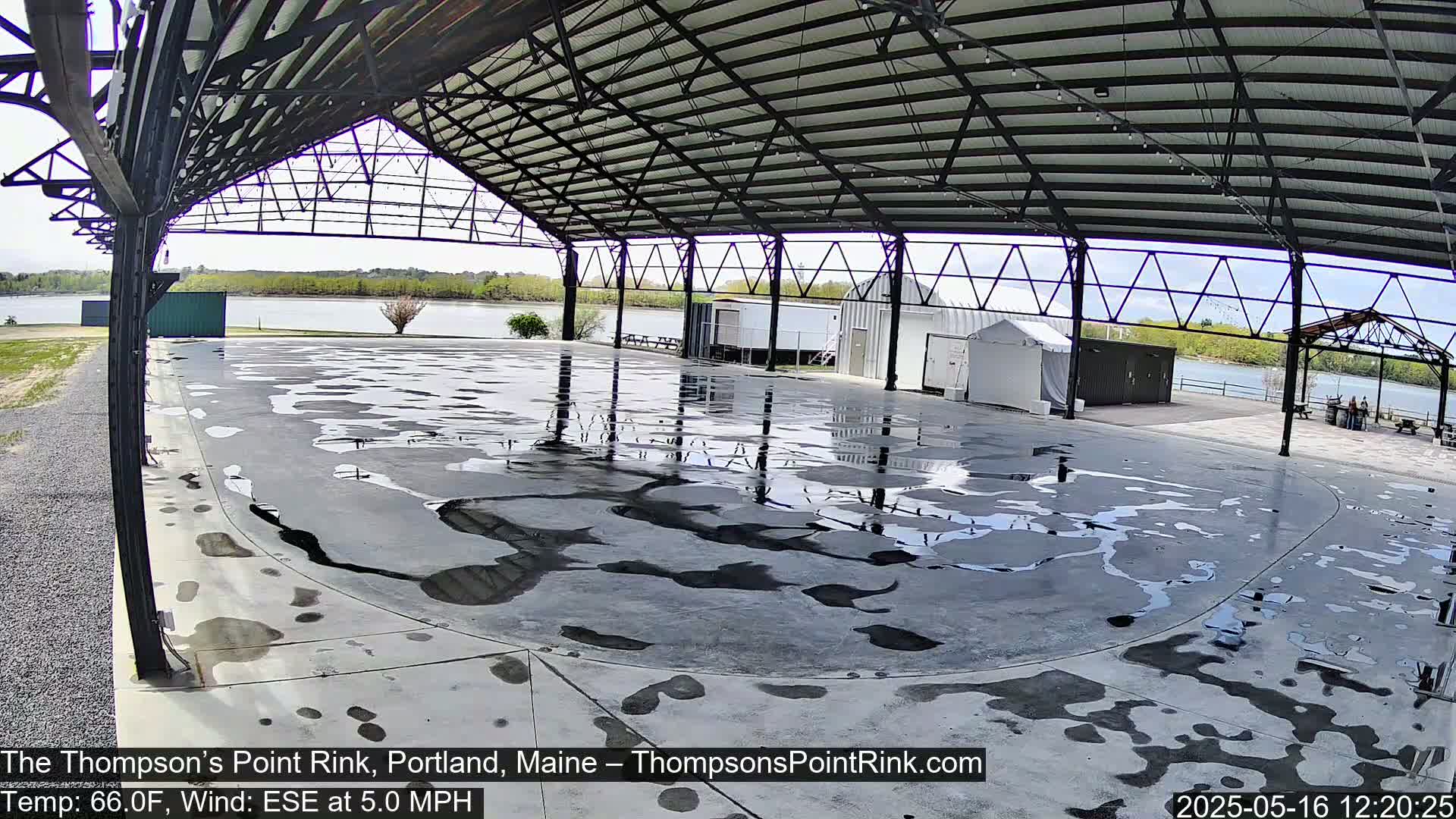 A wet, open-air concrete rink sits under a metal roof structure overlooking a calm body of water on a partly sunny day with a light wind.