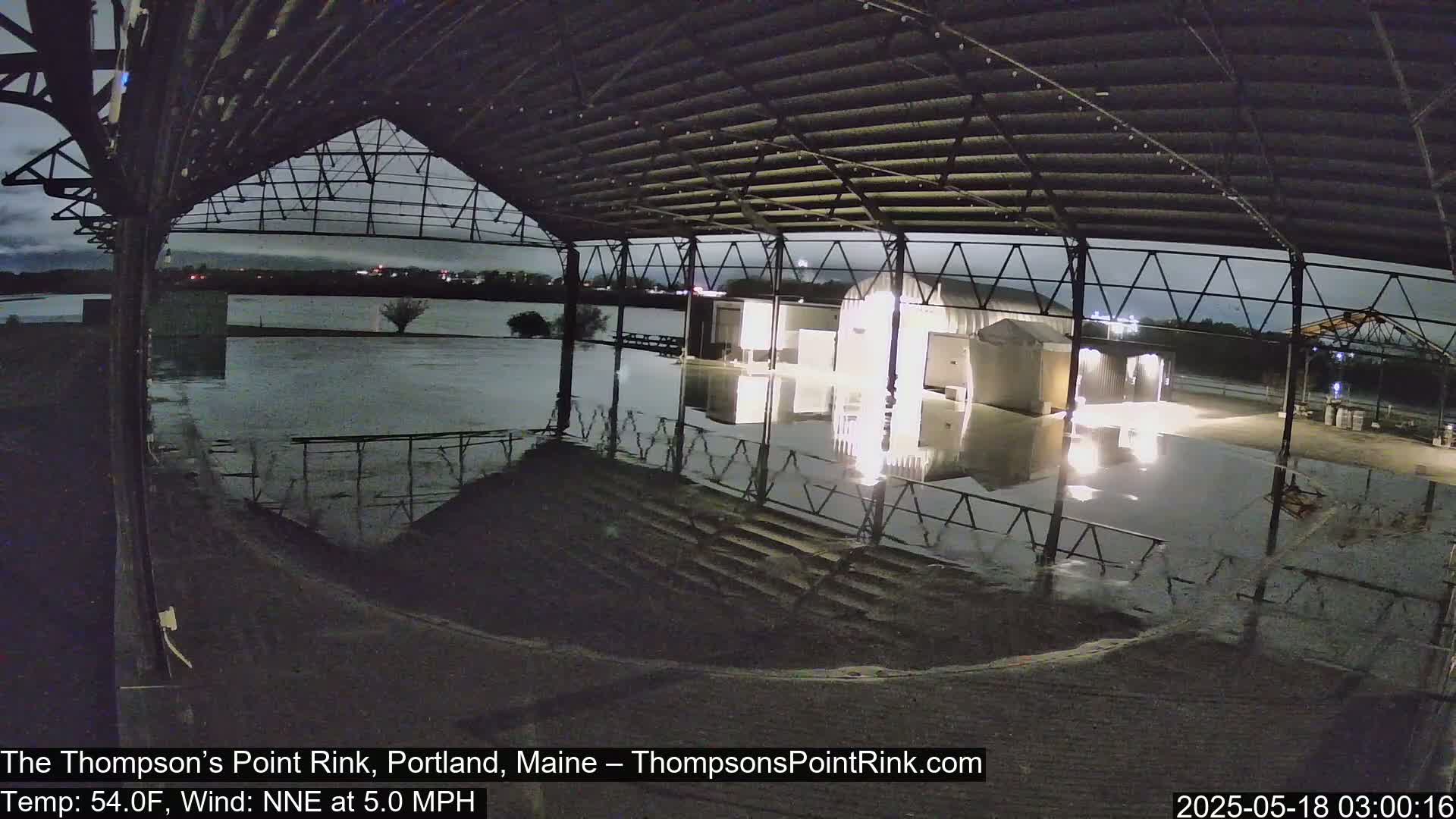 A flooded outdoor rink is viewed from inside a partially enclosed structure at night under a dark, cloudy sky.