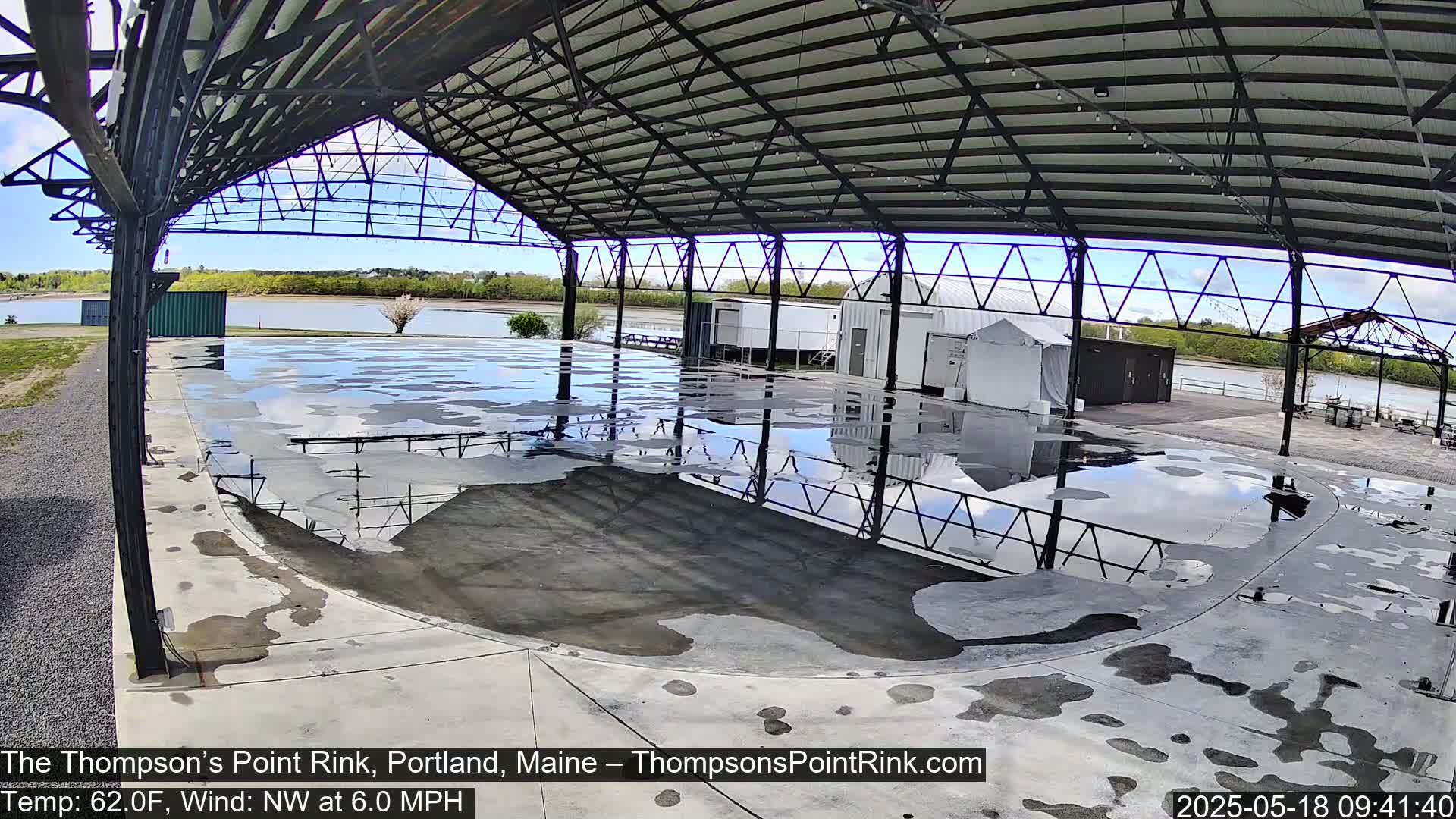 A wet, open-air rink under a metal structure reflects a partly cloudy sky, with a calm body of water visible in the background.