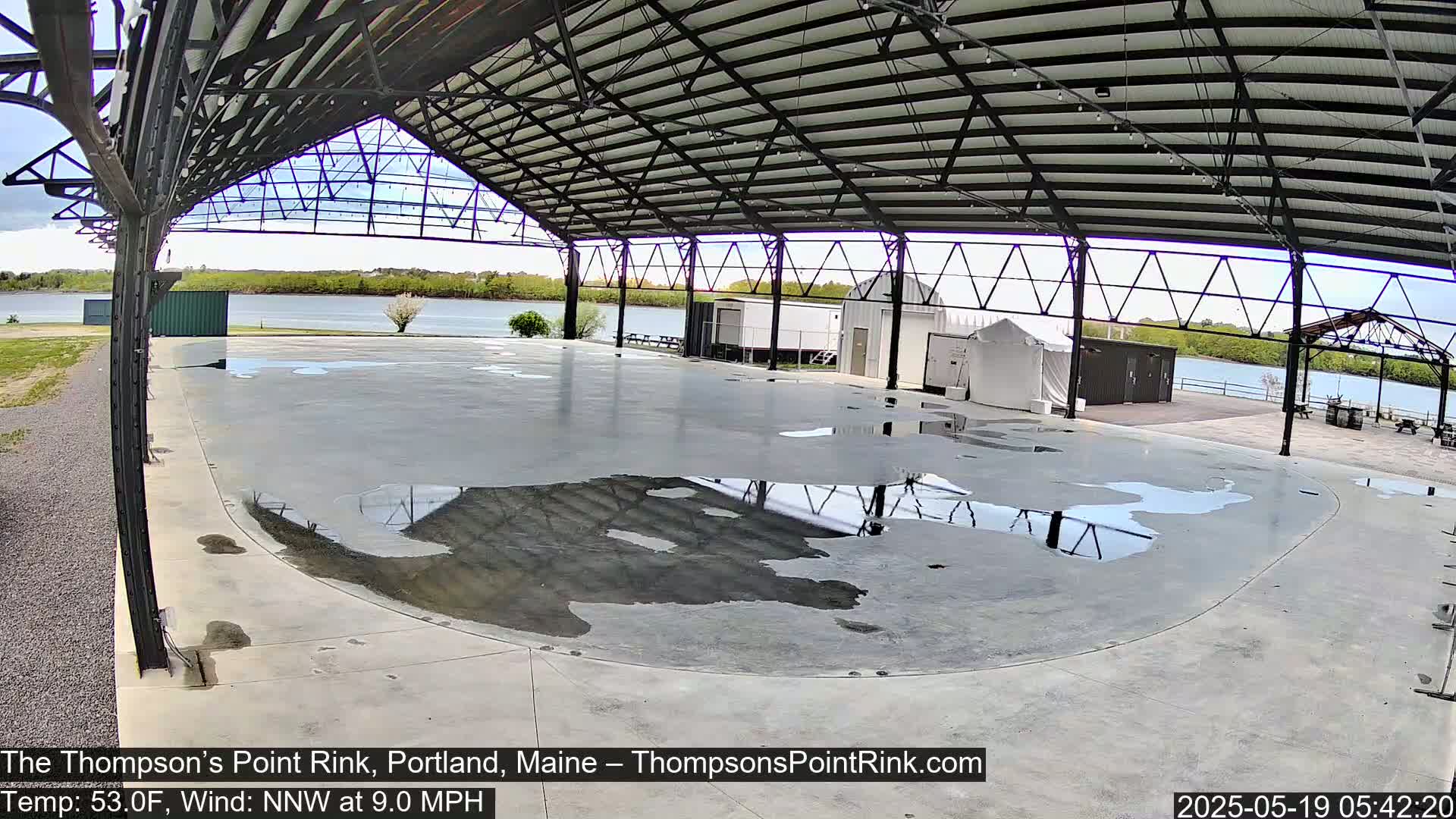 A wet, open-air rink sits under a metal roof structure overlooking a calm body of water on a partly cloudy day.