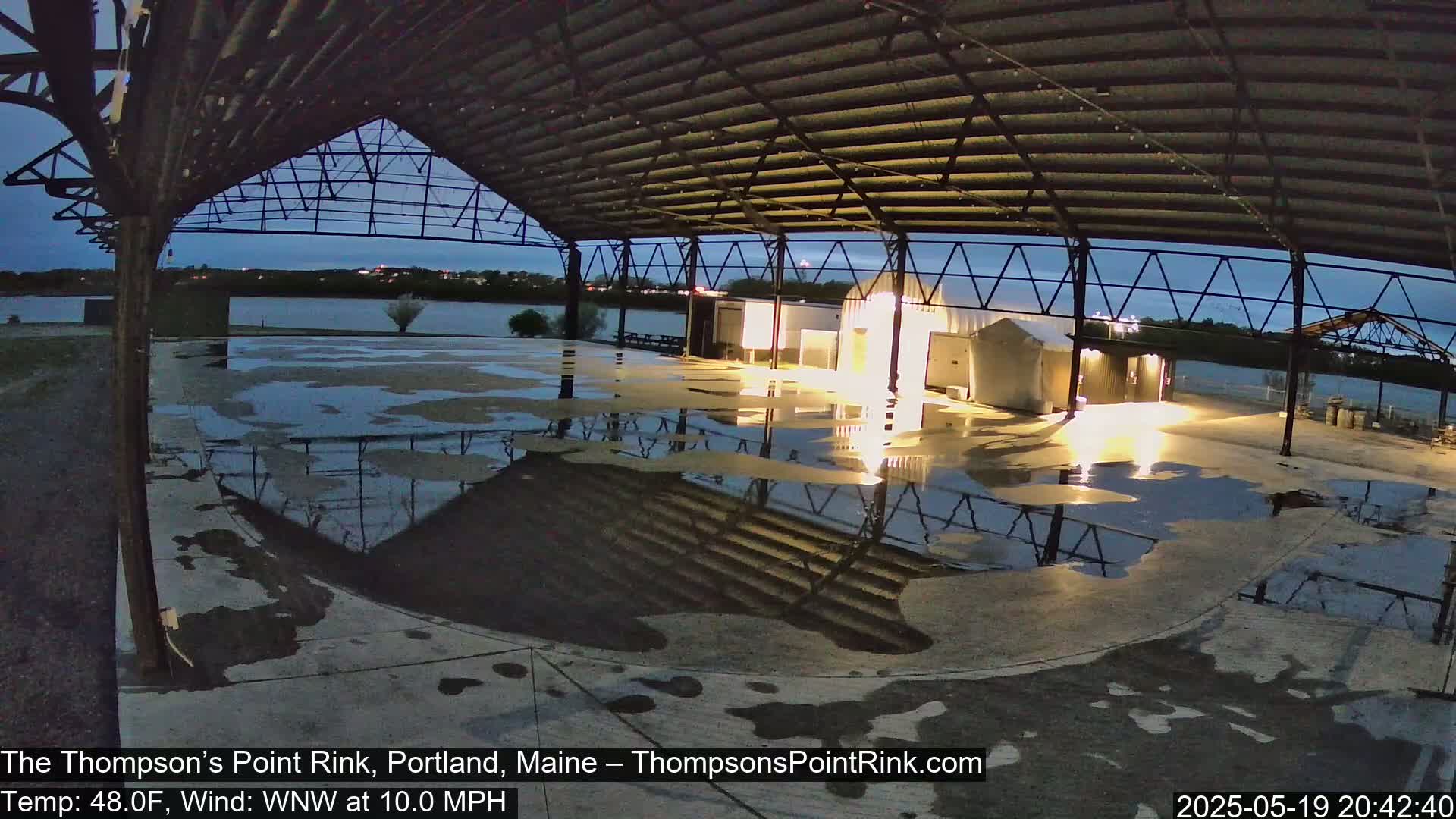 A wet, open-air rink under a metal framework at night, with a calm body of water visible in the background.