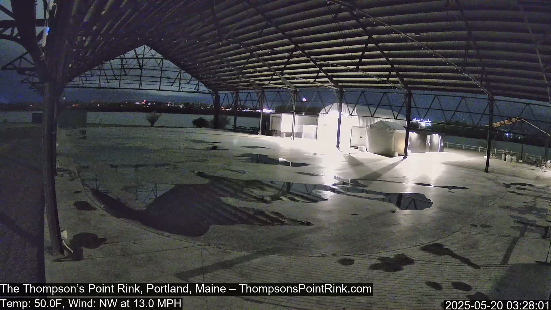 A nighttime view of an empty outdoor rink under a metal roof with puddles on the ground and a northwest wind.