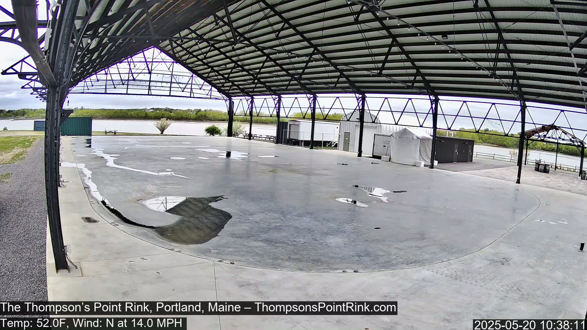 A large, empty, open-air rink with a metal roof is wet from recent rain under an overcast sky.