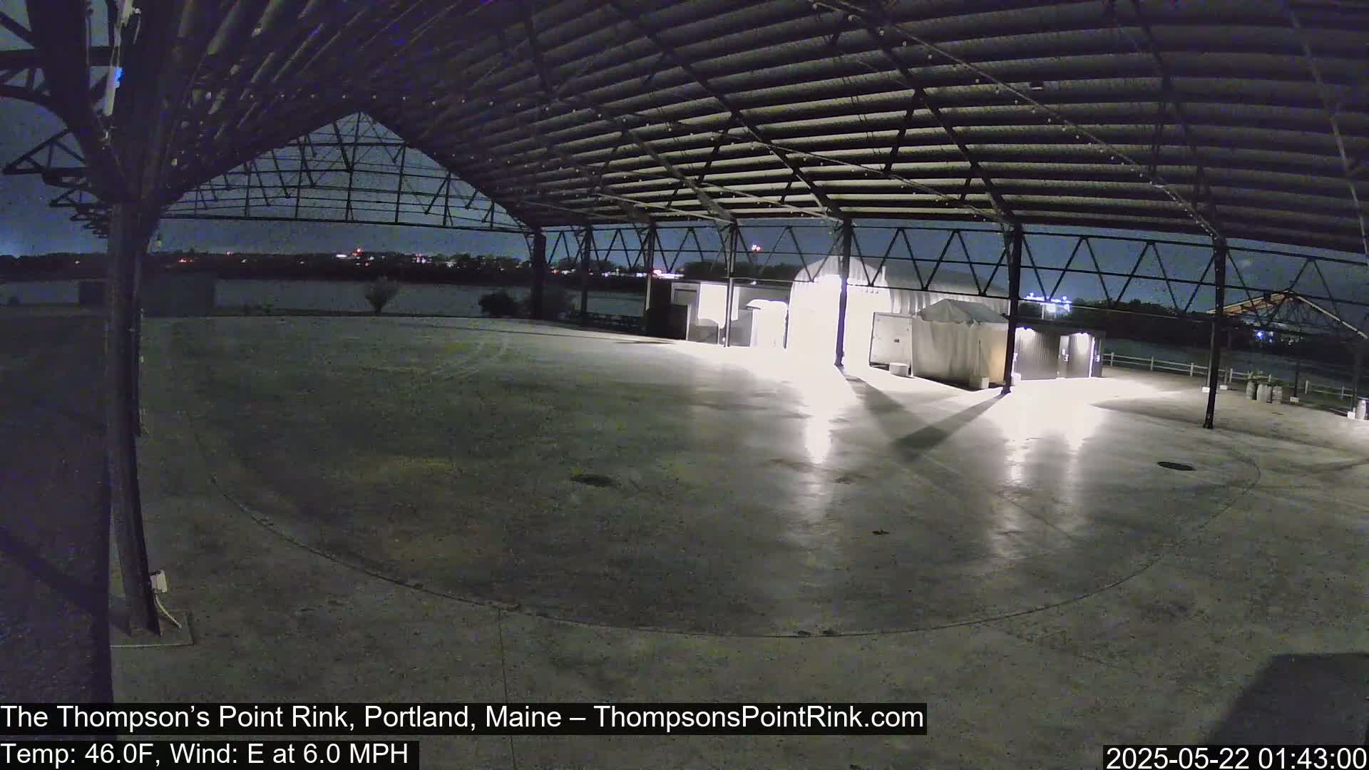 An empty, open-air rink with a metal roof is illuminated at night under a clear sky, with a light east wind.