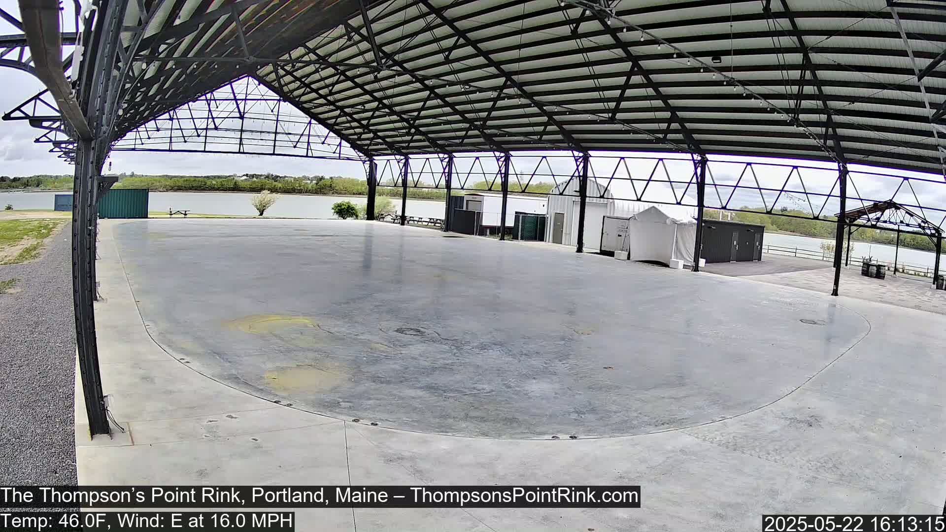 A large, empty, paved rink sits under a metal-framed open-air structure overlooking a calm body of water on an overcast day.