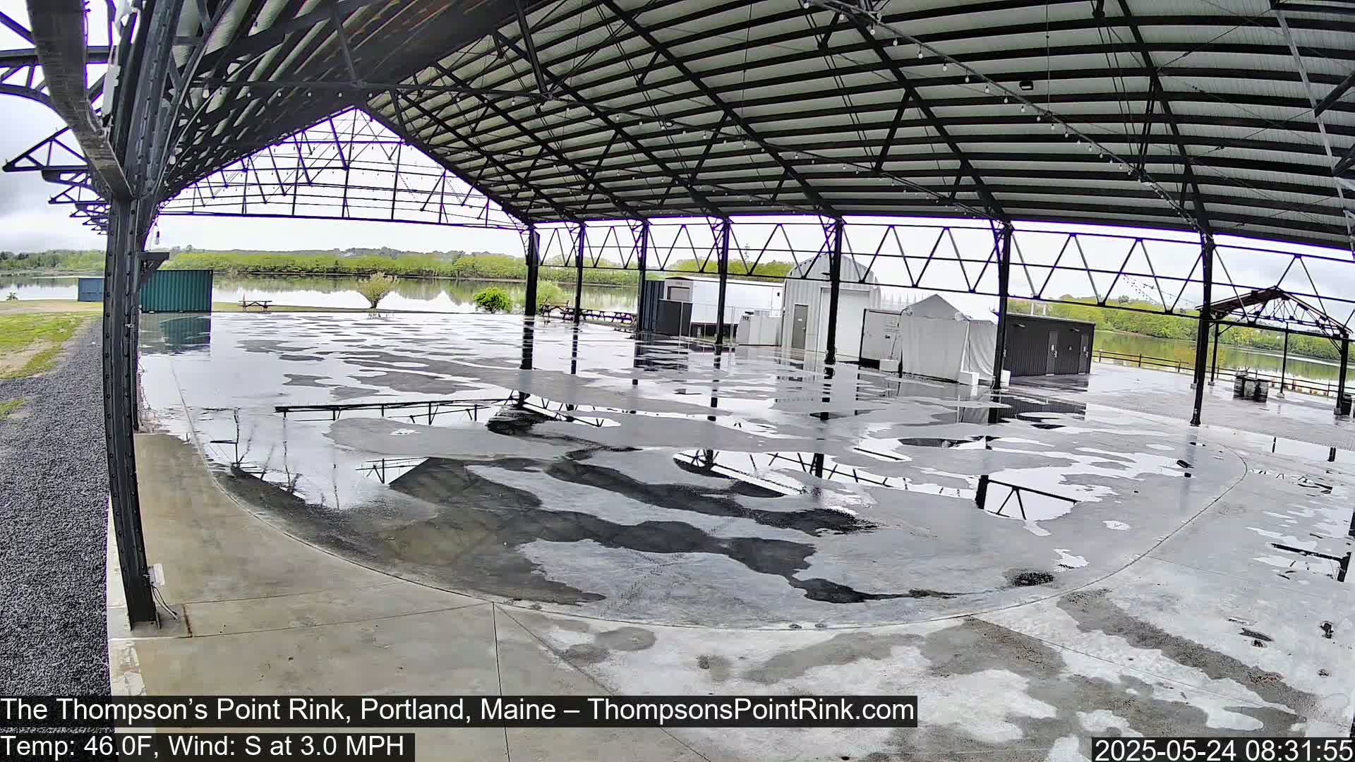 A wet, open-air rink under a metal roof overlooks a calm body of water under an overcast sky.