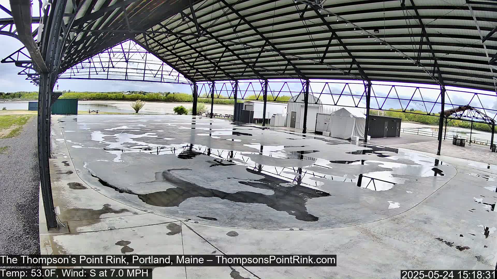A wet, open-air rink under a metal roof with a partly cloudy sky visible beyond.