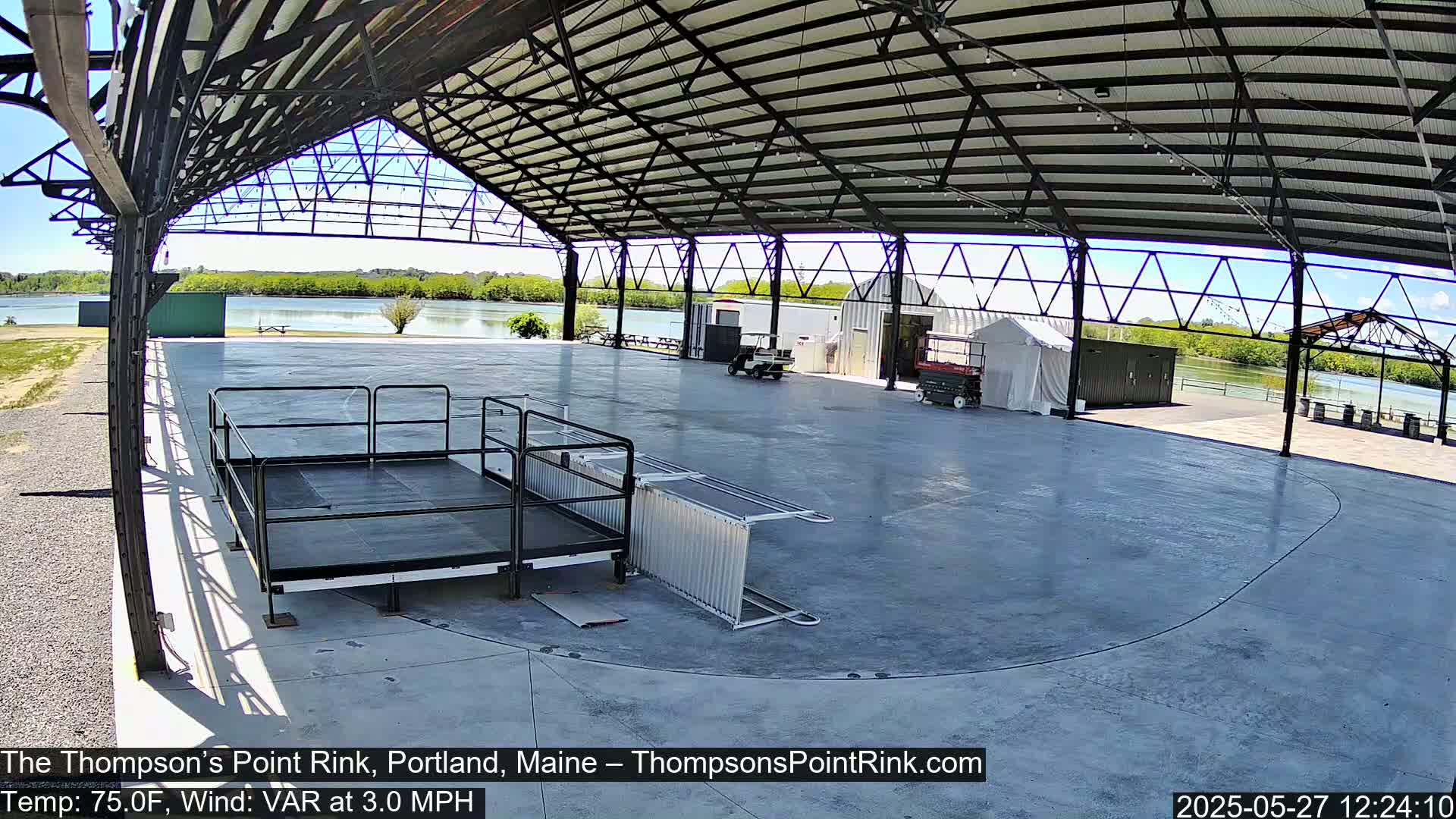 An empty, covered concrete rink sits beside a calm body of water under a sunny sky.