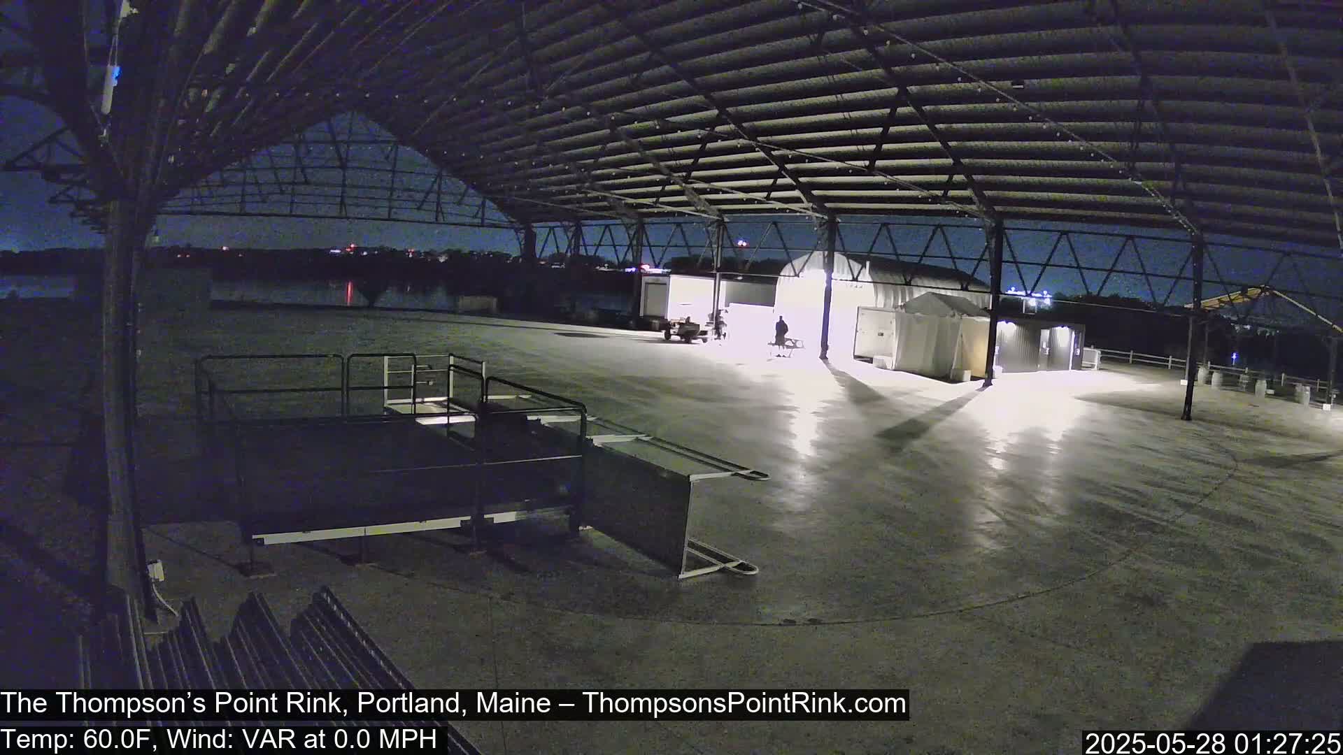 An empty, outdoor rink under a metal roof at night, illuminated by artificial lights, with calm weather.