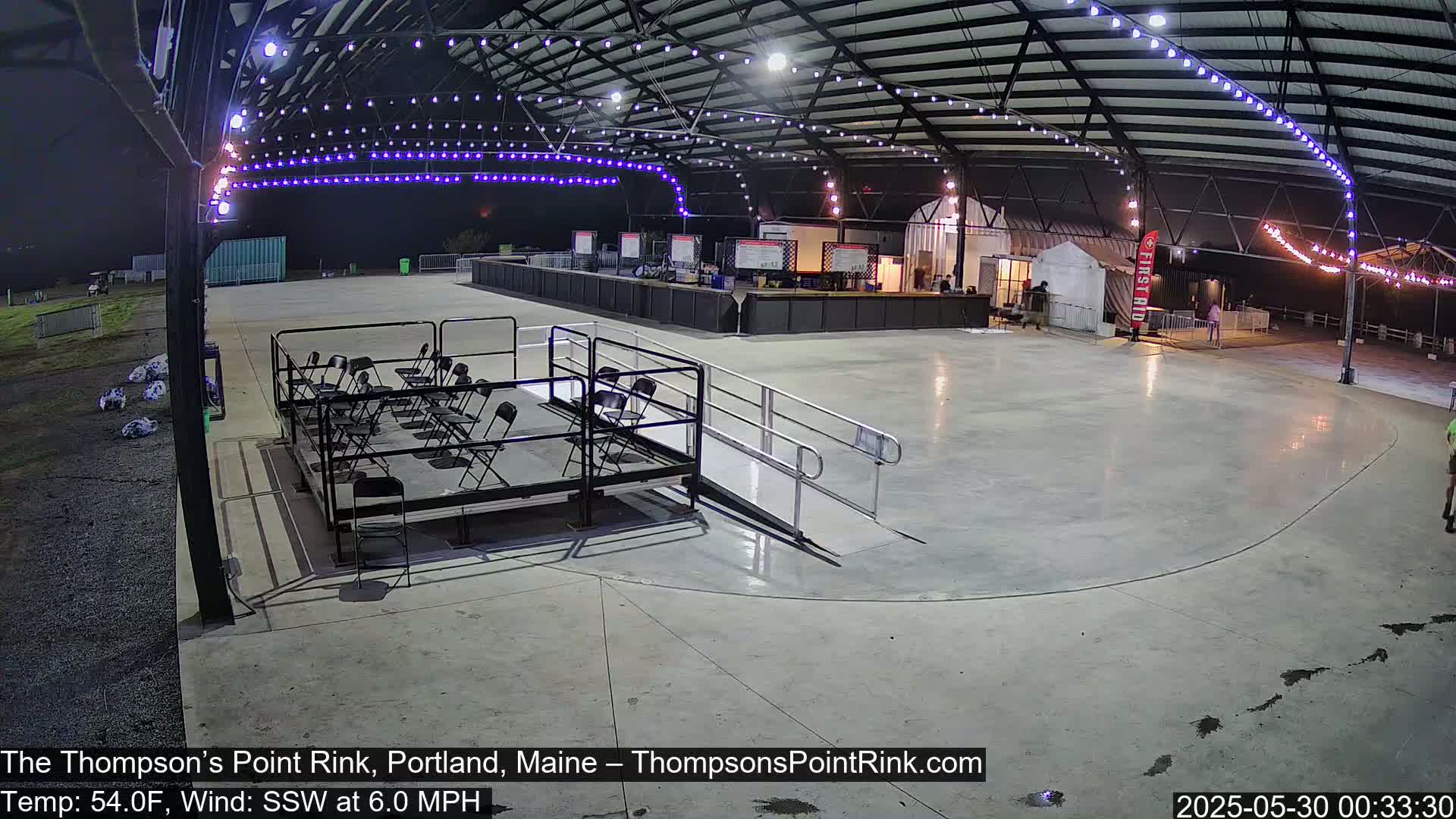 An empty outdoor rink under a metal roof at night with string lights, a few chairs set up, and a ramp leading to the rink, with a calm wind blowing.