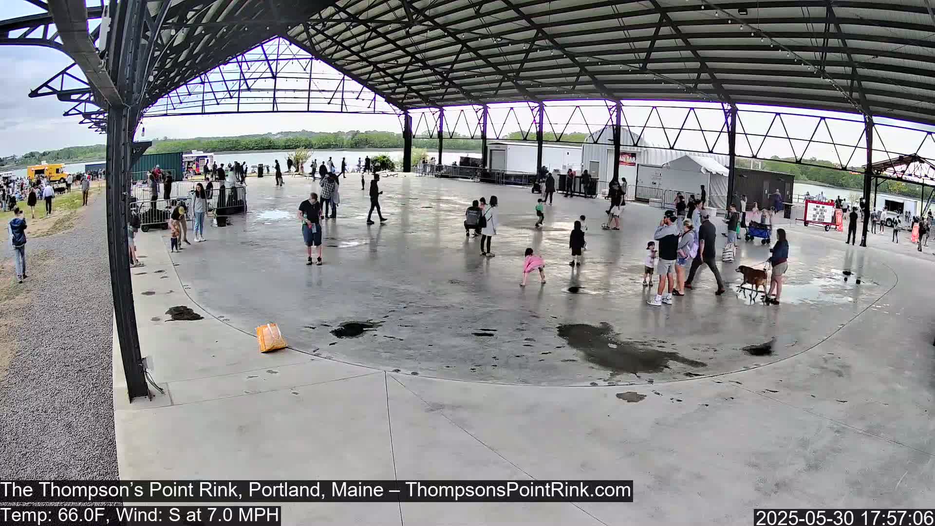 Under a partly cloudy sky, many people are gathered in a large, open-air rink with puddles on the ground.