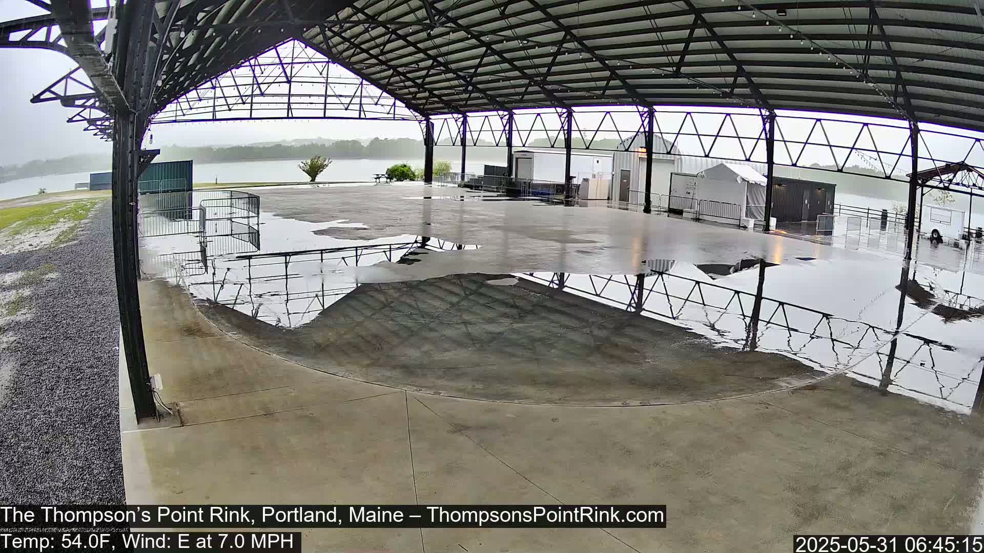 A large, open-air rink under a metal roof is covered in puddles of rainwater under a gray, overcast sky.