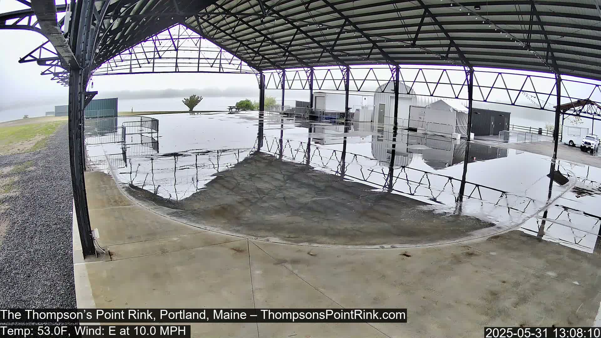 A wet, open-air rink sits under a metal roof on an overcast day.