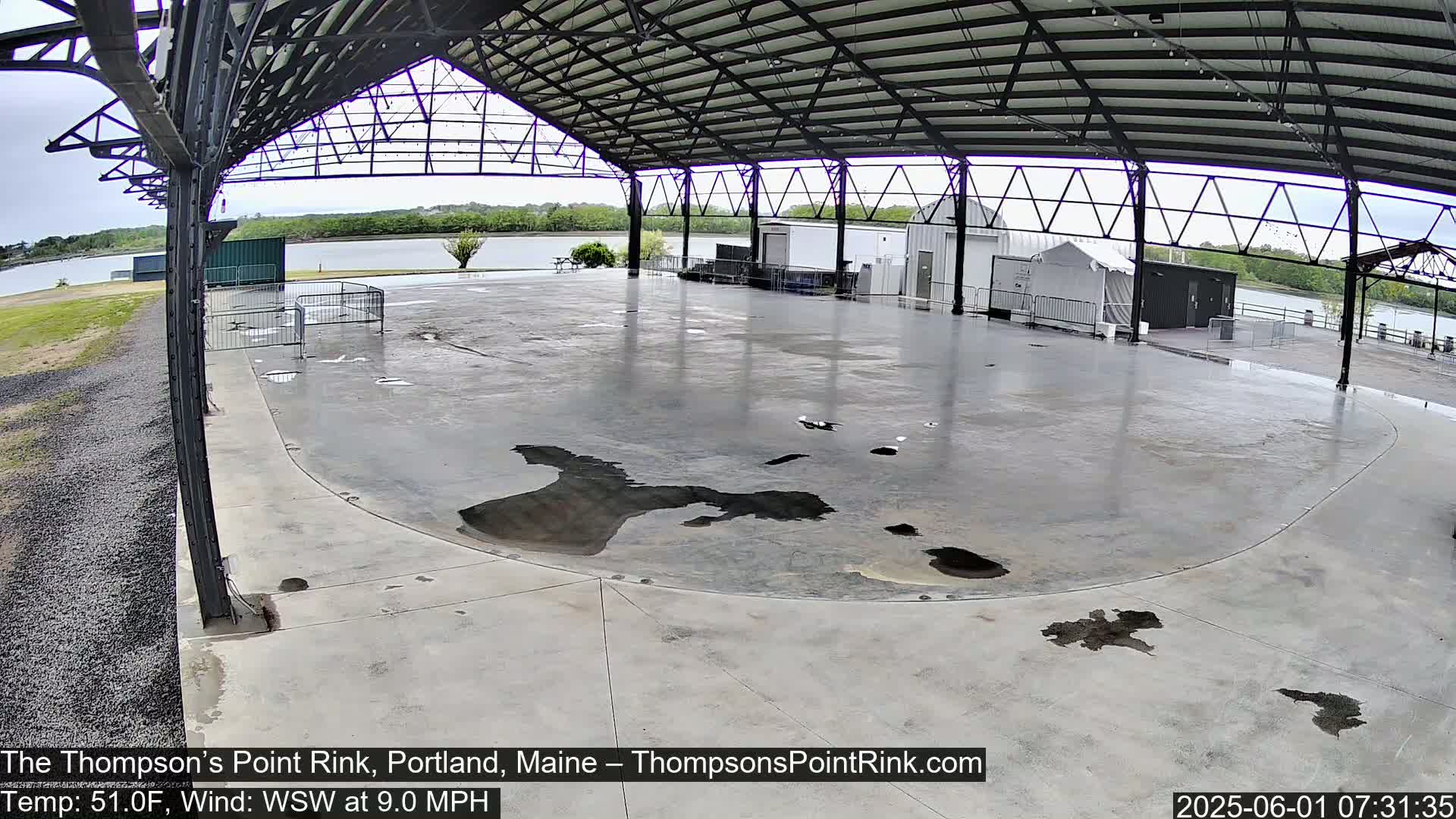 A wet, empty concrete rink sits under a metal roofed structure overlooking a body of water on an overcast day.