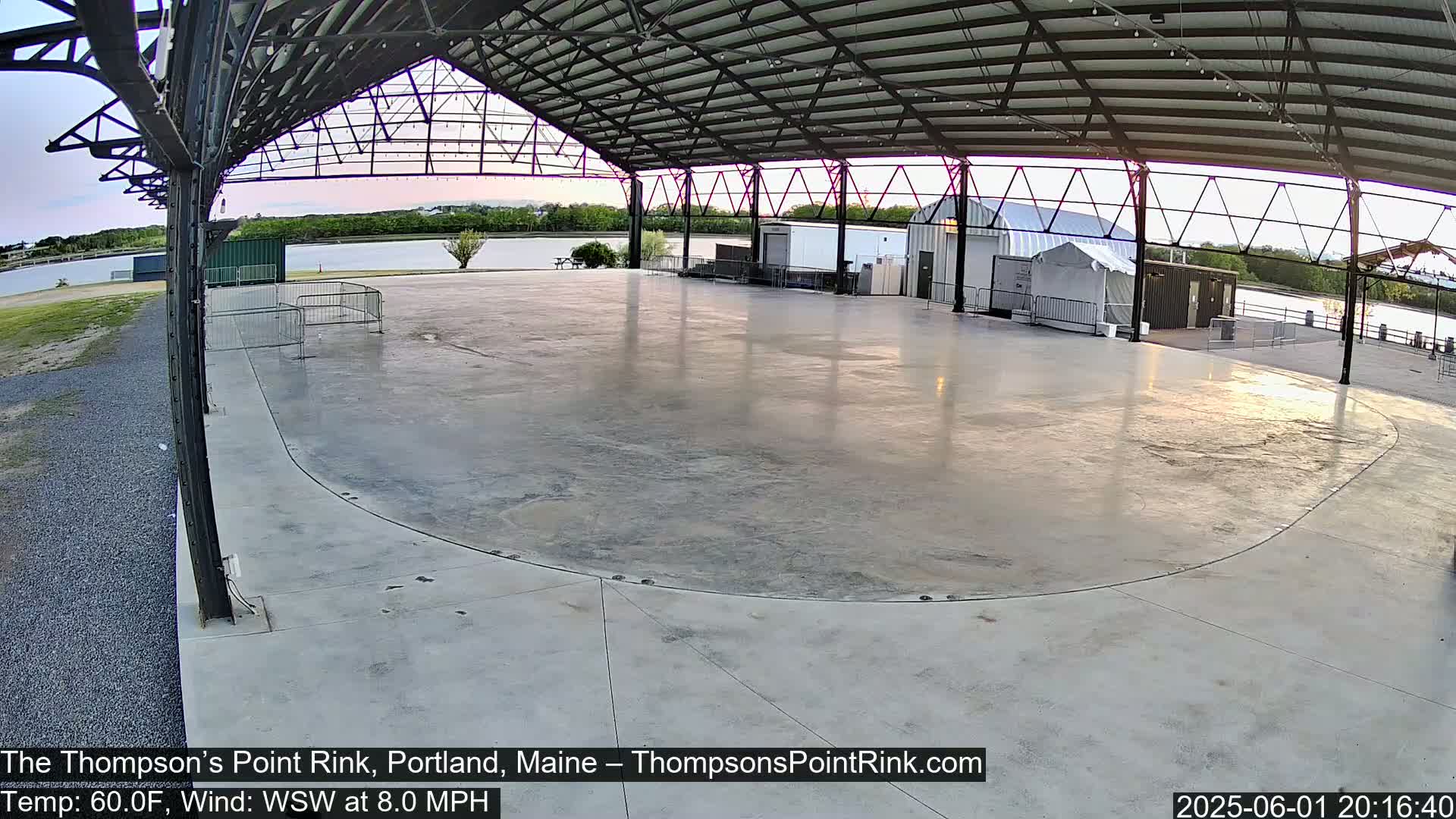 An empty, paved outdoor rink sits under a metal roof structure at dusk, with calm winds.