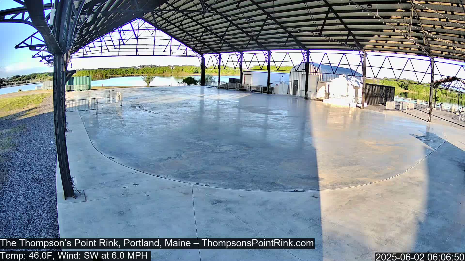 An empty, concrete outdoor rink is situated under a metal-framed shelter, overlooking a calm body of water under a clear sky.